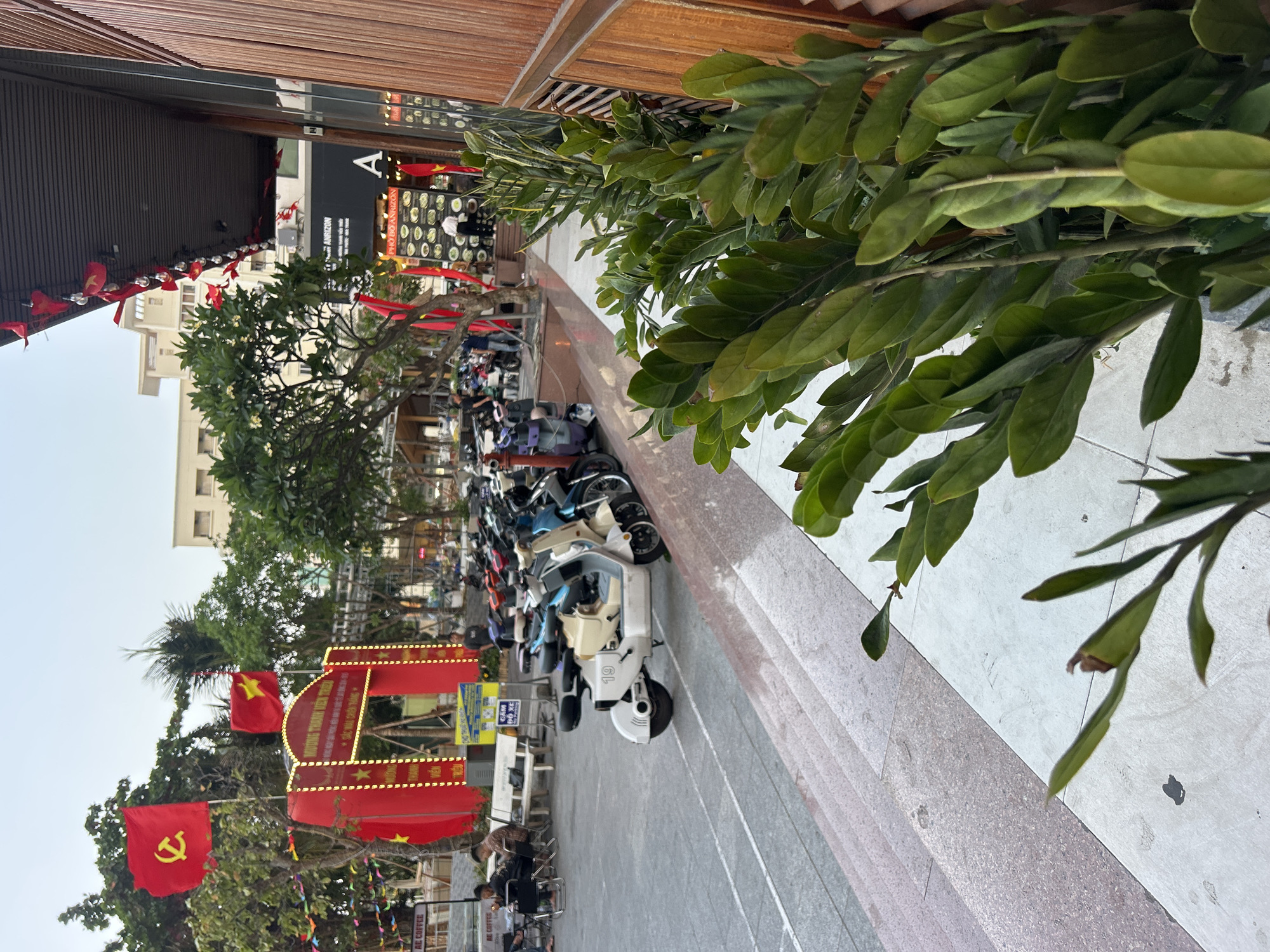 The image captures a street scene in Bắc Nha Trang, Vietnam. Motorbikes are parked along the edge of a sidewalk lined with greenery. Red flags, some bearing a hammer and sickle emblem and others with a star, are prominently displayed, likely in preparation for a celebration or festival. An illuminated red archway or gate is visible, further decorated with flags and lights, suggesting a festive atmosphere. A storefront with "Pho Do Amazon" printed on it is also visible. People can be seen walking and sitting, likely patrons of nearby establishments. The scene is brightly lit, indicating it's daytime.