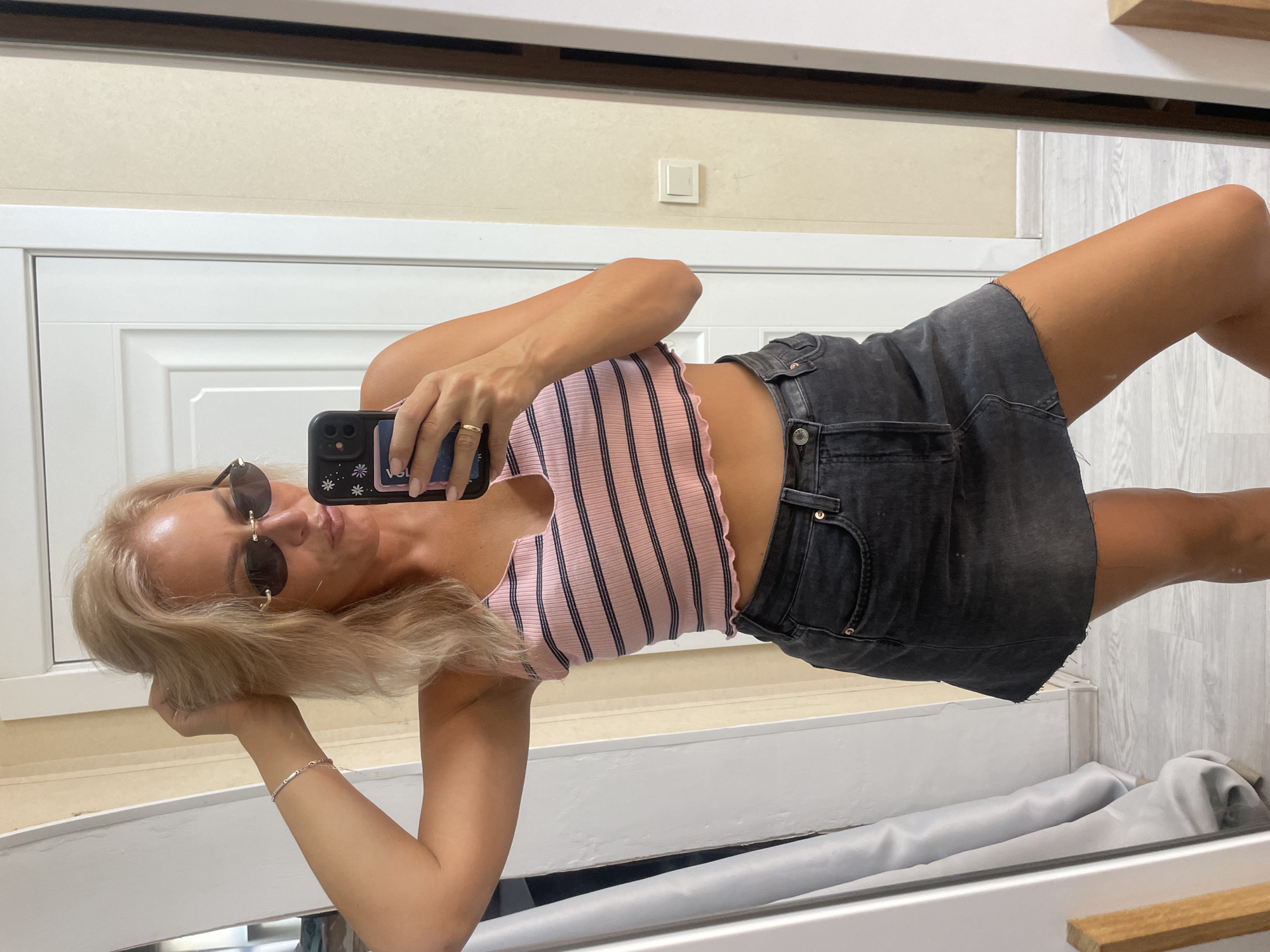 A blonde woman is standing in front of a mirror and taking a selfie. She is wearing a pink and navy striped crop top and a dark denim skirt. She has on sunglasses and a ring on her finger. The room appears to be a bedroom or dressing area, with a white door and a wall with a light switch visible in the background. The floor is wooden. There is no indication of the city of Baranovka, Russia.