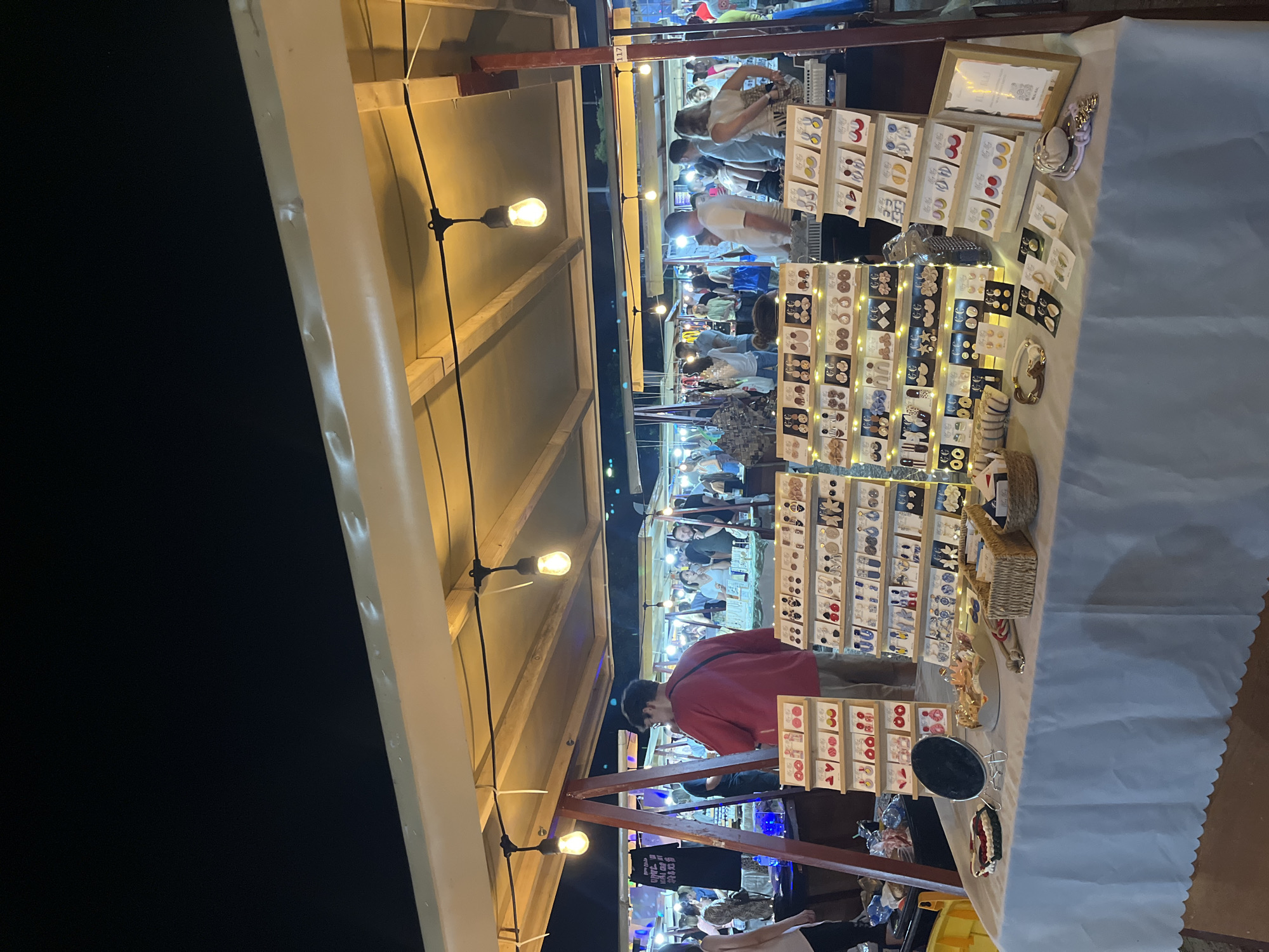 This is a nighttime outdoor market scene, likely at a fair or festival, with a focus on a stall displaying handmade jewelry, particularly earrings. The stall is covered by a light-colored canopy illuminated by strings of warm-toned Edison-style light bulbs. The foreground features a table draped with a white tablecloth, displaying various pairs of earrings mounted on cardboard cards. These earrings come in a variety of styles, colors, and materials, including geometric shapes, floral designs, and studs. There are also some bracelets and other accessories visible on the table.
Behind the table, several racks are filled with more earrings, neatly arranged and lit by small LED lights. A few people are browsing the stall, including a man in a red shirt and a woman examining the jewelry. In the background, the market extends further, with other stalls and a crowd of people visible under more lighting. The overall atmosphere suggests a lively evening shopping experience. The question of this location being in Belgrade, Serbia, is not supported by any visual information in the image.