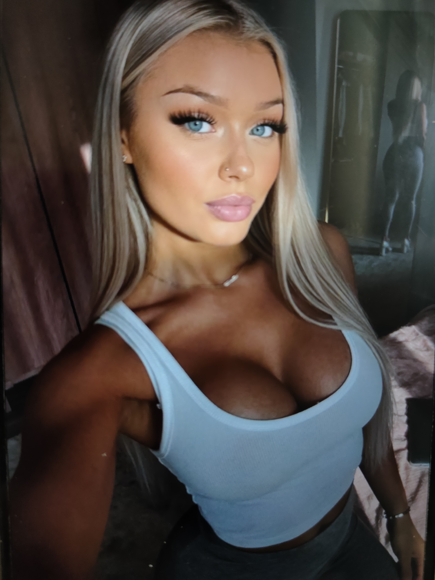 A woman with long blonde hair and blue eyes is looking at the camera. She is wearing a white tank top and grey pants. She has a nose ring and a necklace. She is standing in front of a mirror, and a blurry figure can be seen in the reflection. The background is dark and indistinct, with some pink fabric visible on the left side of the image.