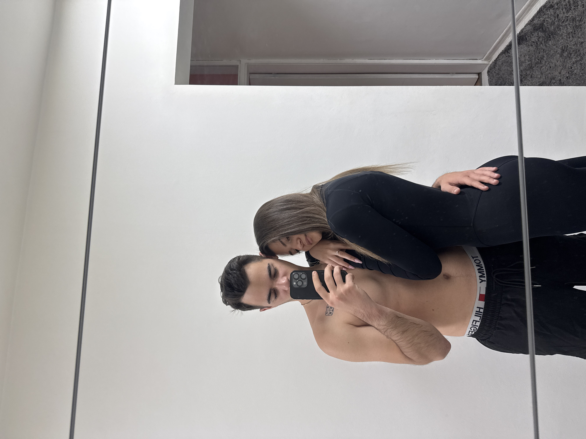 This image captures a mirror selfie of a young couple indoors. The photograph is oriented horizontally, showing the man and woman leaning against each other. The man is shirtless, wearing black athletic shorts with the mirrored "TOMMY HILFIGER" brand name visible on the waistband. He holds a black smartphone, possibly an iPhone, in front of him to take the picture, partially obscuring his face. A small tattoo is visible on his left shoulder area.
Behind him, the woman leans her head affectionately on his back or shoulder, looking towards the camera with a subtle smile. She has long brown hair and wears a long-sleeved black top and black leggings. A ring is visible on her left ring finger.
The setting, reflected in the mirror, shows a minimalist interior space with white walls. Above the main white wall, parts of a ceiling with crown molding, doorframes, and a textured gray surface (possibly carpet or a rug) are visible, indicating an indoor environment. The image conveys a sense of closeness and affection between the couple. The provided location context for this scene is Sofia, Bulgaria.