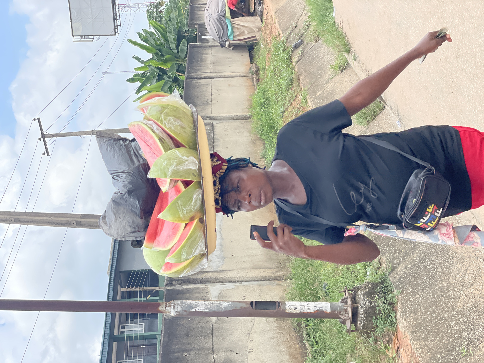 In Fegge, Nigeria, a woman balances a tray of sliced watermelons on her head. She is wearing a black t-shirt and red skirt, with a small black bag slung across her body. In her right hand, she holds a mobile phone, and in her left, she holds a wad of cash. The woman is walking along a street, with a concrete wall and vegetation to her right, and a utility pole to her left. The sky is cloudy. In the background, there are buildings and trees.