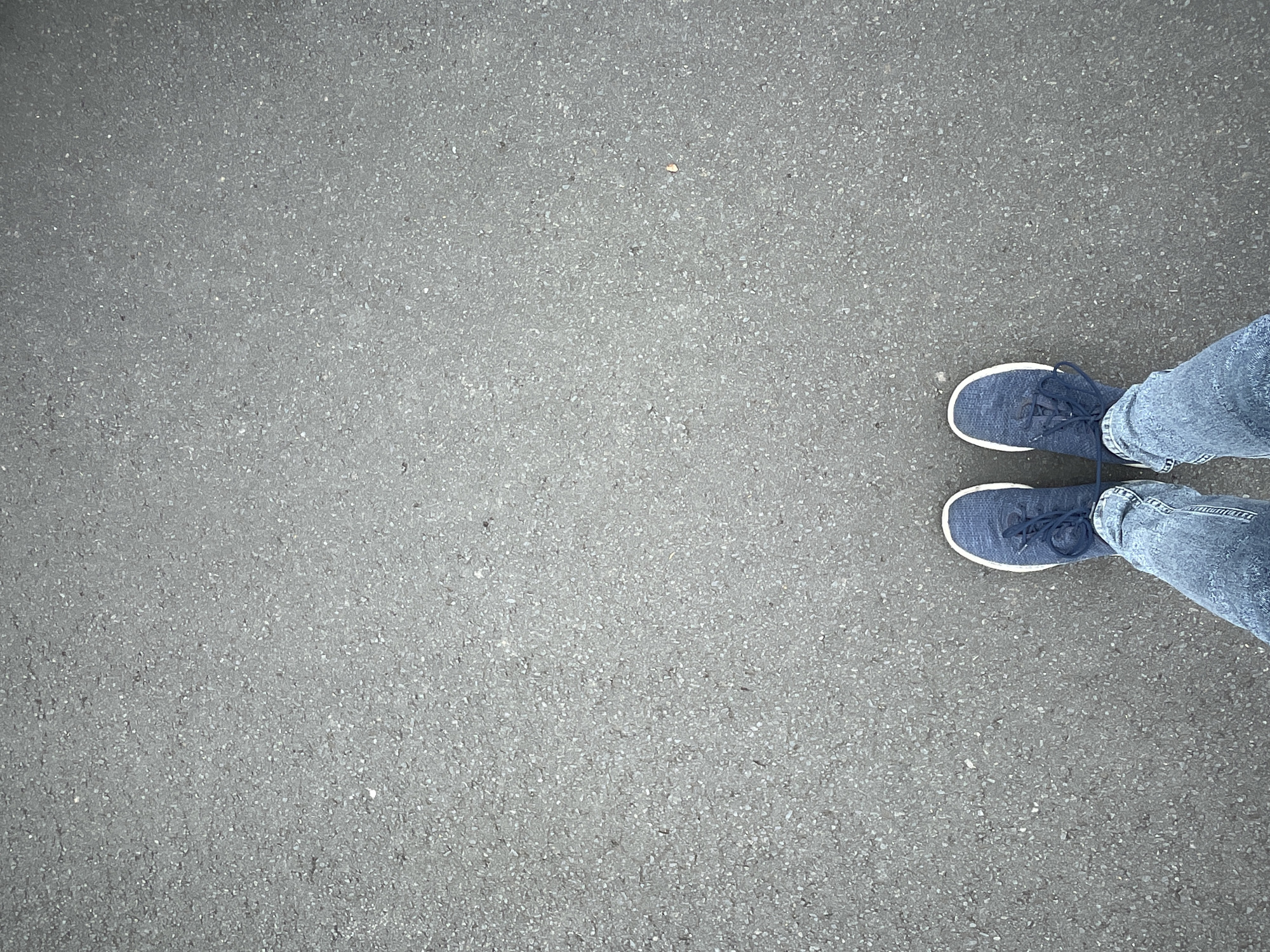 The image shows a top-down view of a person's feet and lower legs standing on asphalt. The person is wearing blue sneakers with white soles and laces, and blue jeans. The asphalt is a dark gray color with small white and gray aggregate. The scene is outdoors, with no other objects or people visible. The location is specified as Rückershausen, Germany.