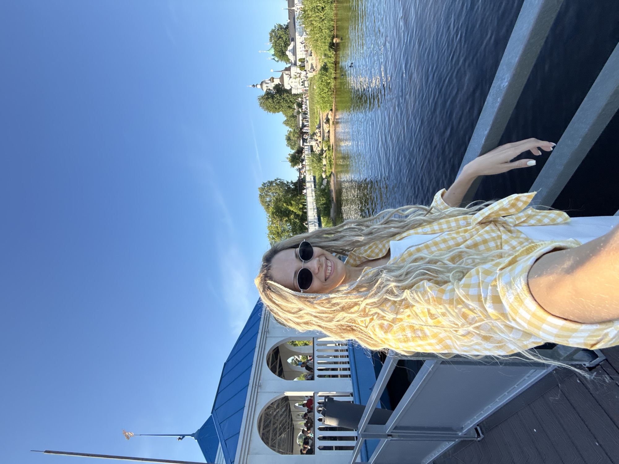 A young fair-skinned woman is centered in a selfie taken on a wooden pier under a clear blue sky. She has long, wavy, blonde hair with dreadlock-like braids or extensions, wears black oval sunglasses, and displays a wide smile revealing teeth. Her attire consists of a white top under a yellow and white gingham plaid button-up shirt, tied at the waist. Her right arm extends forward, holding the camera, while her left hand rests on a grey metal railing with white-painted fingernails.
To the woman's immediate left, a white gazebo-like structure with a blue metal roof is partially visible, featuring open arches where several individuals are seated. The pier is constructed of dark wooden planks. In front of the woman, a body of dark blue water is present, with two ducks visible on its surface in the midground.
Across the water, a treed and landscaped bank with green foliage and sandy patches is visible. A white bridge with railings spans across this area, and multiple people are walking on the bank and the bridge. In the far background, a prominent complex of white buildings with multiple dark and green domes and spires is visible, consistent with Russian Orthodox architecture, resembling a monastery or church. The scene is brightly lit by natural sunlight, indicating daytime. The location is specified as the city of Rusanovo, Russia.