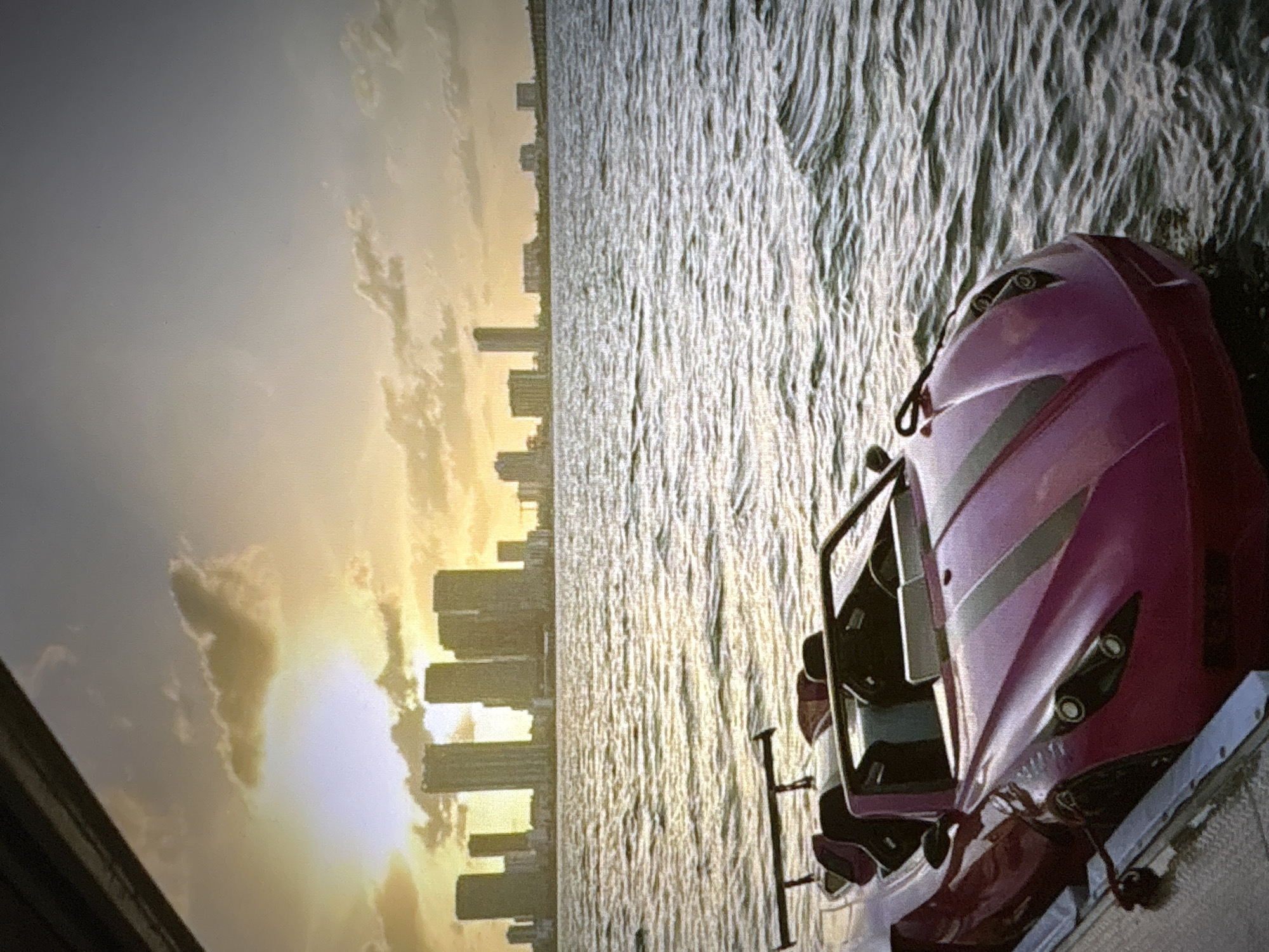 The image shows a pink sports car with a spoiler, docked on a platform next to a body of water. The water has moderate ripples, reflecting the sky. In the background, a cityscape with numerous buildings is visible across the water, silhouetted against a sunset sky with dramatic clouds. The sun is setting, casting a warm, golden light on the scene. The image composition focuses on the car in the foreground, with the city and sunset serving as the backdrop. The photograph appears to be taken from a slightly elevated perspective. The location is not Pori, Finland; it is a city with a skyline typical of a coastal metropolis, likely in a warmer climate.