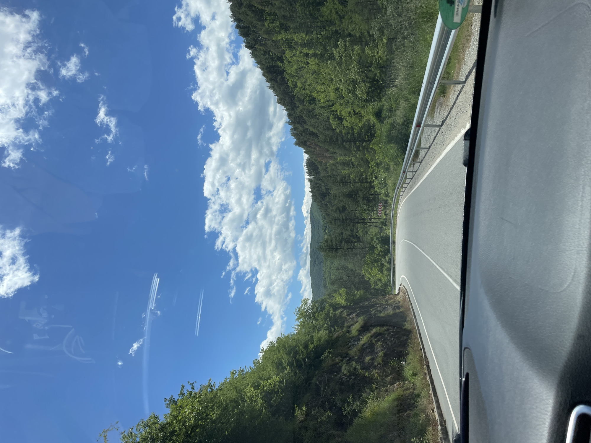 The image depicts a view from a vehicle traveling on a paved road winding through a forested area near Startsevo, Bulgaria. A metal guardrail runs along the right side of the road. The sky is visible to the left, featuring scattered clouds. The dashboard of the vehicle is visible in the lower right corner.
