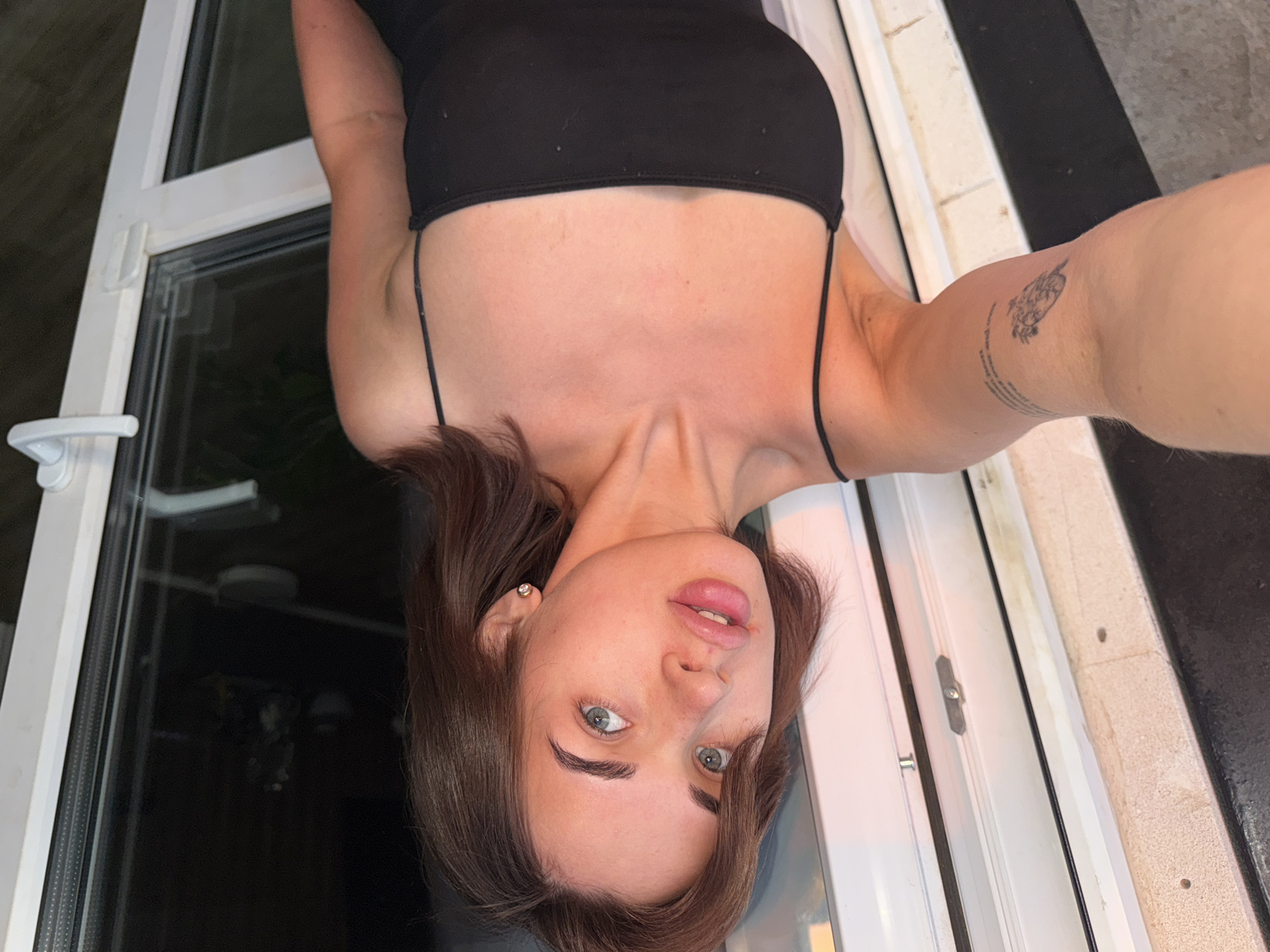A young woman is positioned near a window, taking a selfie. She has brown hair, fair skin, and is wearing a black top with spaghetti straps. A tattoo is visible on her upper arm. The window frame is white, and a view through the glass shows an obscured dark outdoor scene. It is not possible to confirm the location is Лобаново, Russia, from the image.