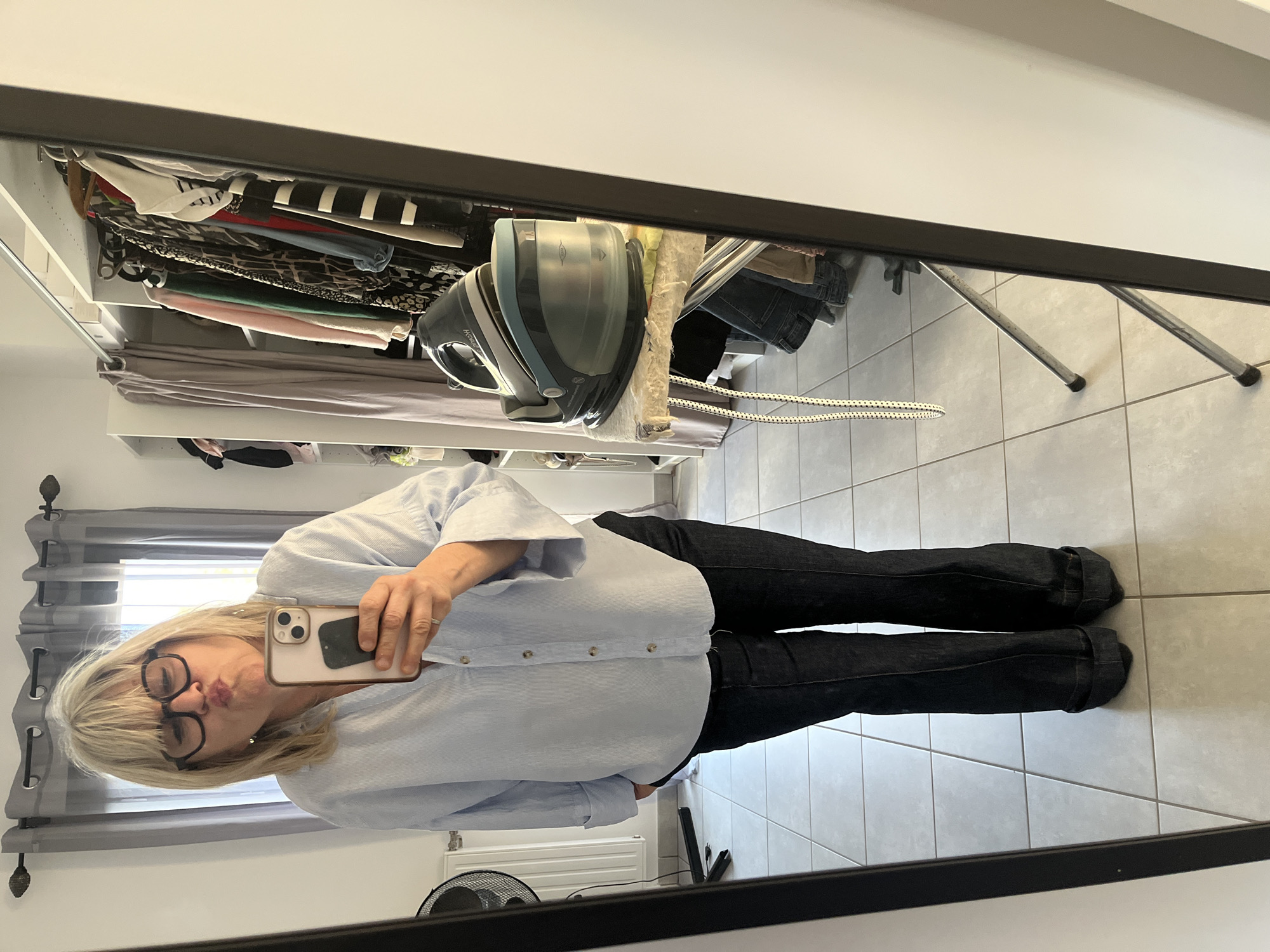 A woman stands in front of a mirror, taking a selfie with a smartphone. She is wearing glasses, a light blue button-down shirt, and dark jeans. The room appears to be a bedroom or dressing area, with a closet containing clothing and an ironing board visible in the background. The floor is tiled. There's a radiator near the bottom of the mirror frame. The location is Chambœuf, France.