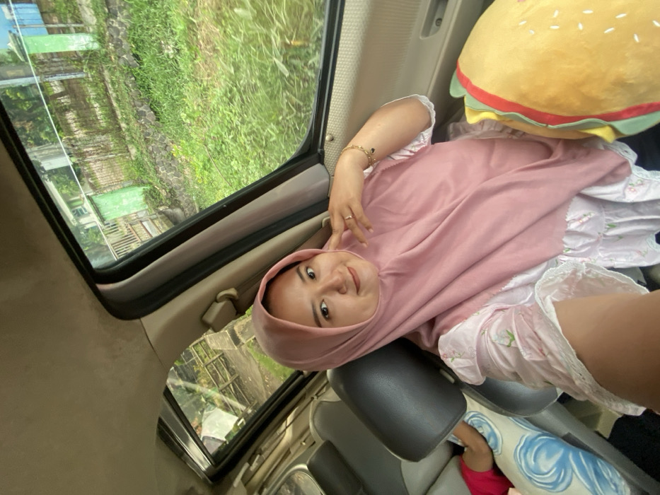 Inside a vehicle with an open sunroof, a young woman wearing a pink hijab and floral dress leans back against a car seat. She is smiling and holding up two fingers in a peace sign. Outside the sunroof, trees and buildings are visible. A large, plush hamburger toy sits beside her. Her feet are visible near a blue pillow and a red shoe. The angle suggests the photo was taken from above.