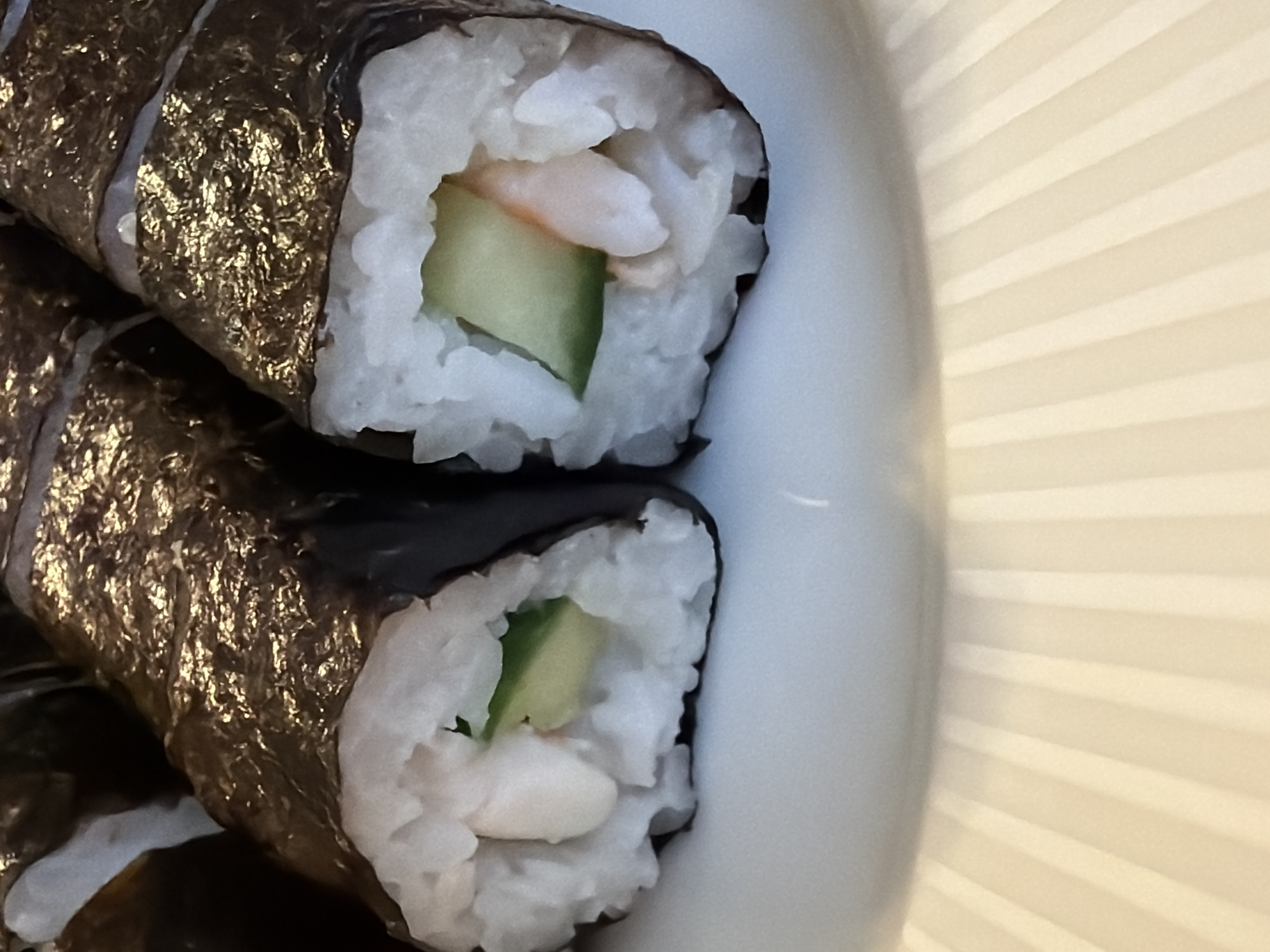 The image shows a close-up of two pieces of sushi rolls on a white plate. The sushi rolls consist of white rice, a dark seaweed wrap (nori), and fillings of cucumber and what appears to be shrimp or a similar seafood item. The plate has a ribbed design. The location is Unknown, Russia. There are no people visible in the image.
