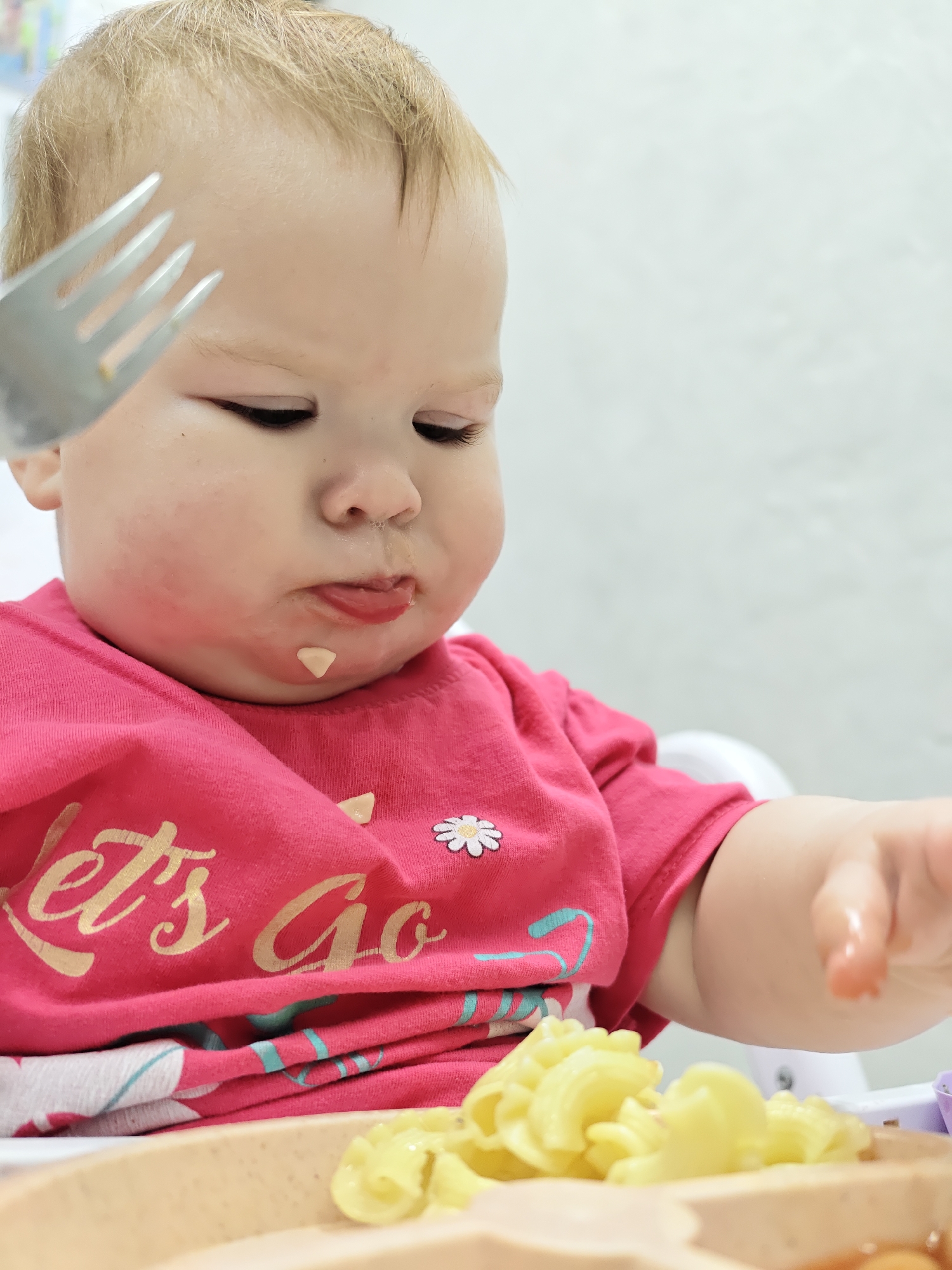 In an indoor setting, a baby sits in a high chair, dressed in a pink t-shirt with the words "Let's Go" printed on it and a small white flower. The baby has blonde hair, and there is food residue on their face. A fork is being held near the baby's face, and a plate of pasta is in front of them. The background is a plain, light-colored wall. The location is Chaikovsky, Russia.