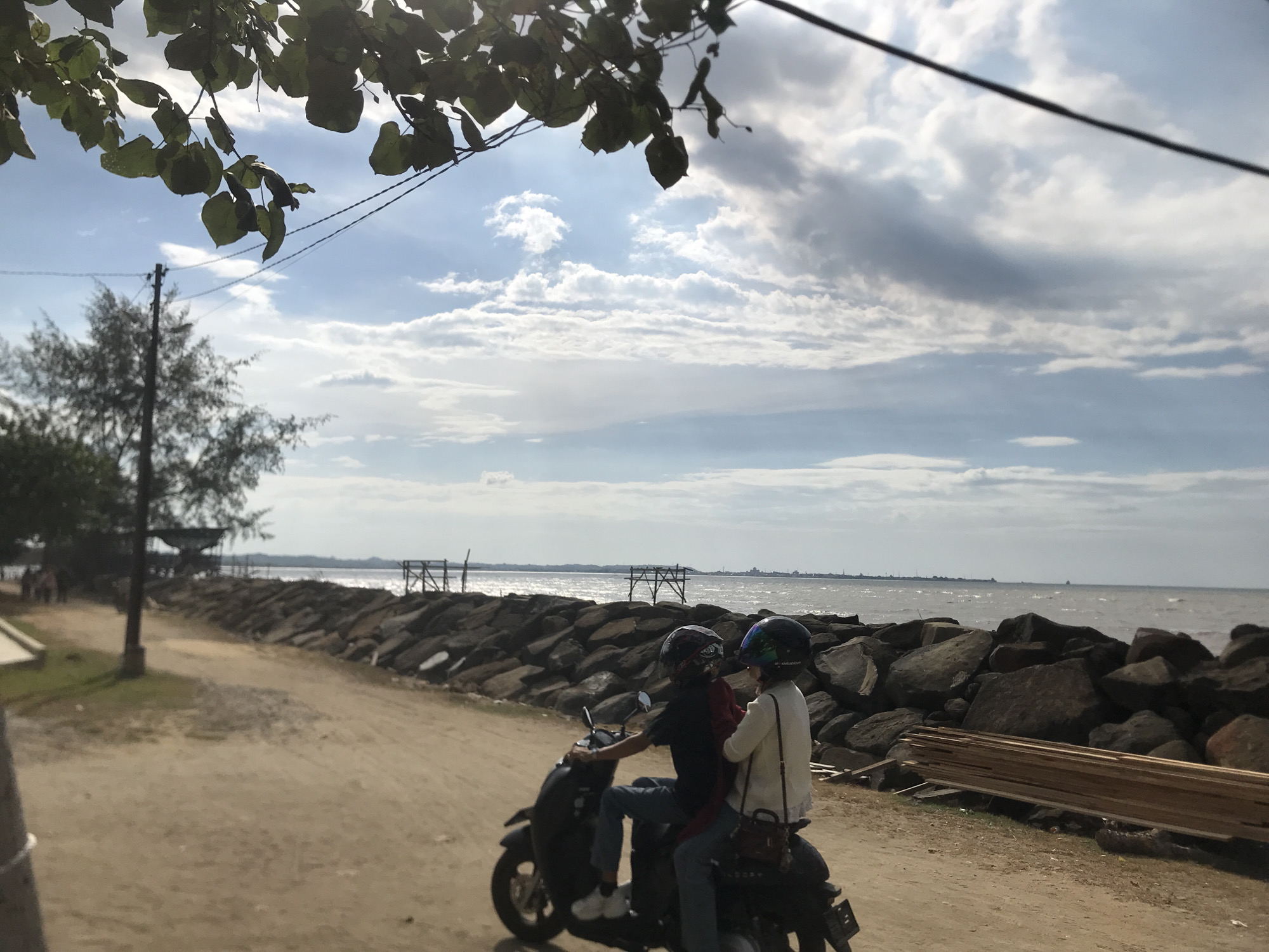 A medium shot depicts two individuals riding a black scooter on a dirt road in Lancok, Indonesia. The road is parallel to a shoreline protected by a breakwater of large gray rocks. To the left, a dirt pathway leads past a light pole and under a small, covered structure near a tree line. Scattered lumber lies alongside the breakwater.
The scooter is occupied by a driver wearing a dark shirt, blue jeans, white shoes, and a dark helmet. The passenger wears a light-colored jacket, jeans, and a dark helmet. Both appear to be facing away from the viewer, towards the horizon.
Beyond the breakwater, the ocean meets a cloudy sky, with a distant cityscape visible on the horizon. The composition is partially framed by leafy branches and a power line in the upper portion of the image.