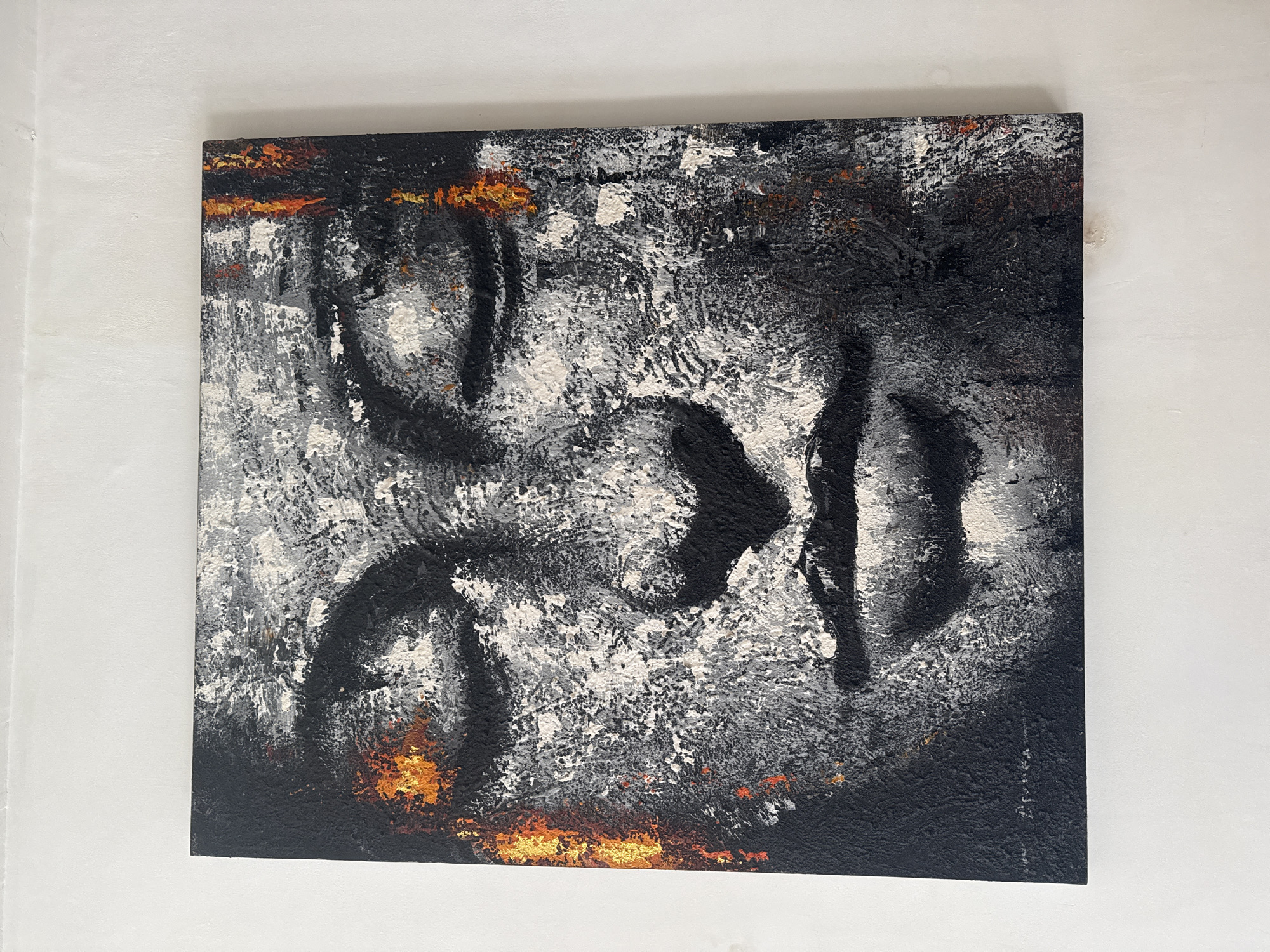 A square abstract painting hangs on a white wall. The painting features heavy textures and layers of black, white, grey, and small accents of orange paint. The shapes within the painting are indistinct and appear to be broad, gestural strokes. There is no indication of people, objects, actions, or location information relating to North Kuta, Indonesia in the image.