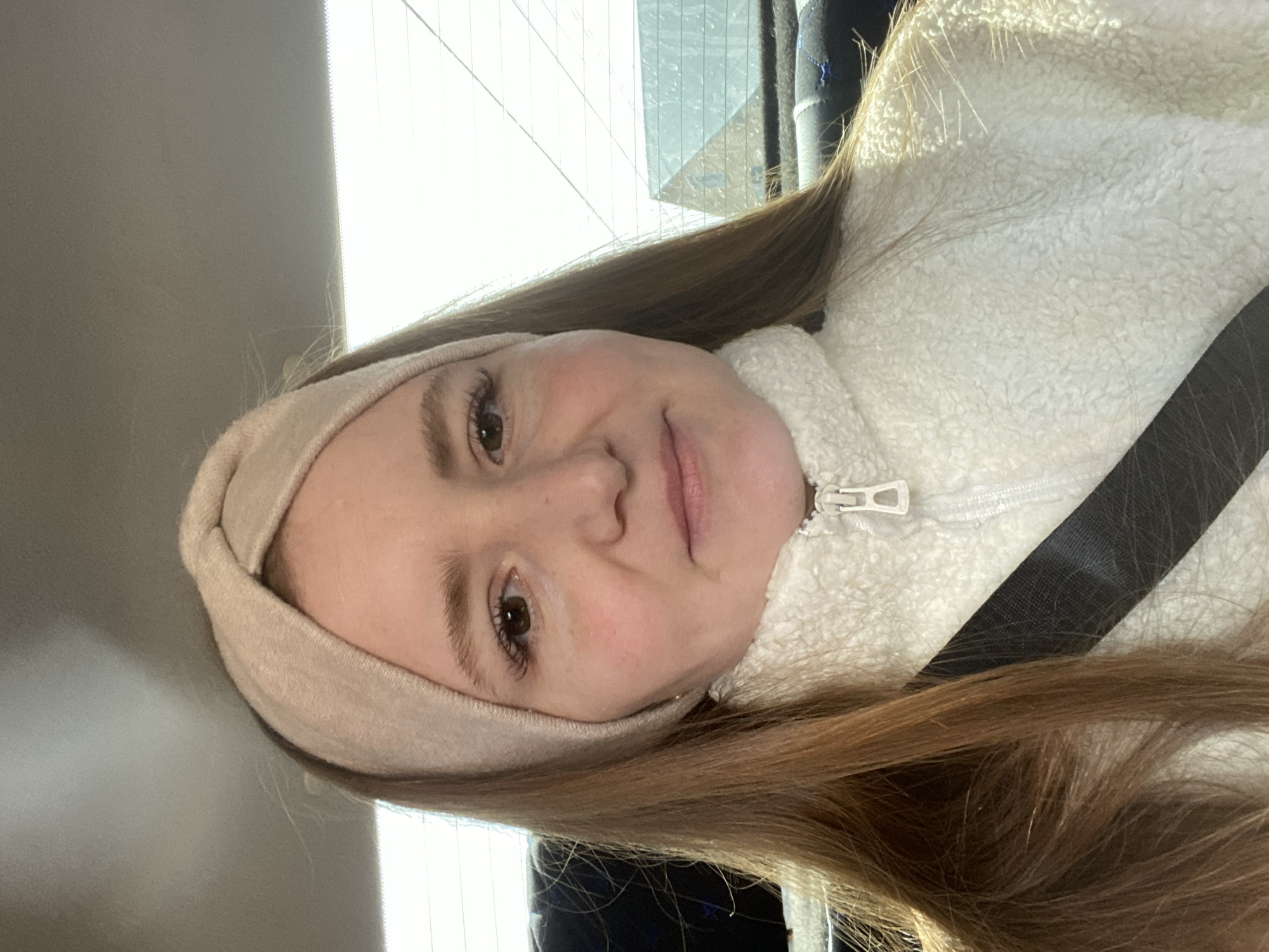 A young woman is facing the camera, wearing a beige headband and a white fleece zip-up jacket, with a black seatbelt visible across her body. The background appears to show a window, likely in a car, suggesting an indoor setting possibly within Chaikovsky, Russia, though no definitive landmarks or signage confirm the specific location. The primary subject is the woman, and she is wearing a seat belt. The environment seems to be a vehicle's interior.