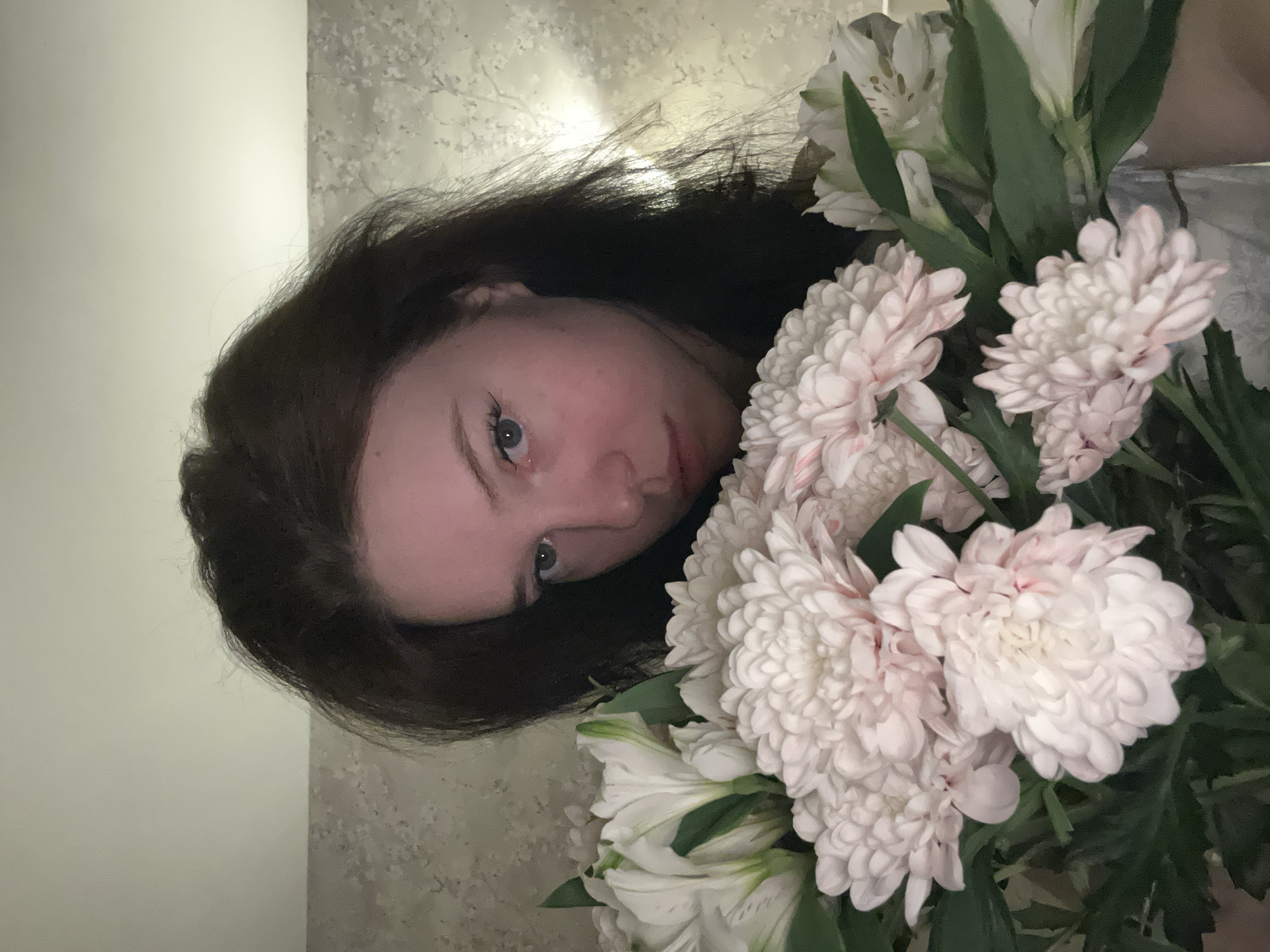 A young woman with dark hair and fair skin is partially visible, her face angled towards the camera. Her eyes are blue. She is holding a bouquet of flowers, including white chrysanthemums and lilies. The background consists of a plain light-colored wall and a patterned surface. It is unknown whether the location is in the city of Chaikovsky, Russia.