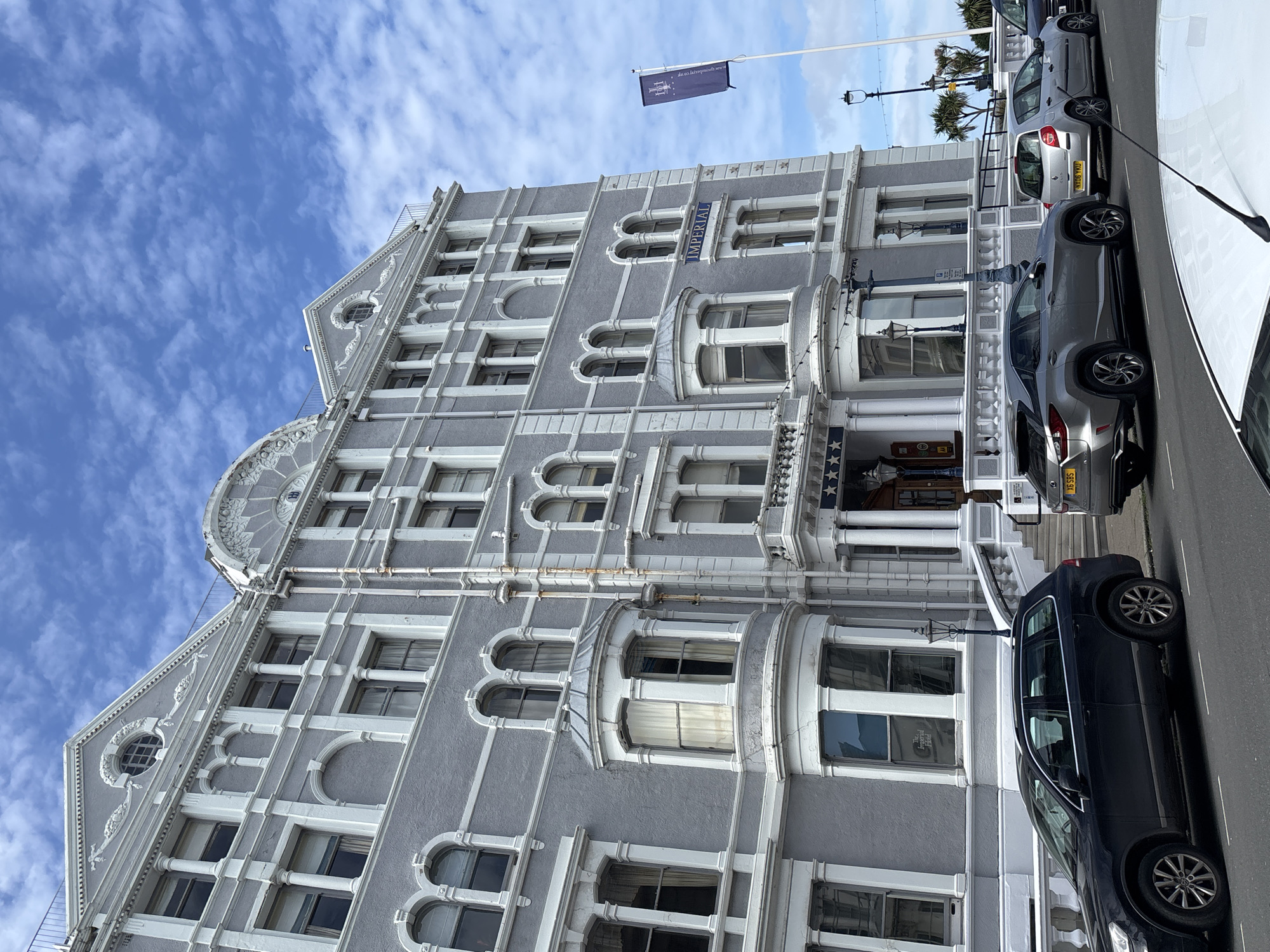 The image shows the exterior of the Imperial Hotel in Llandudno, United Kingdom. The hotel is a large, gray building with white architectural details, including arched windows, decorative molding, and columns. A flag is visible on the roof of the building. Cars are parked along the street in front of the hotel. The sky is blue with scattered clouds.