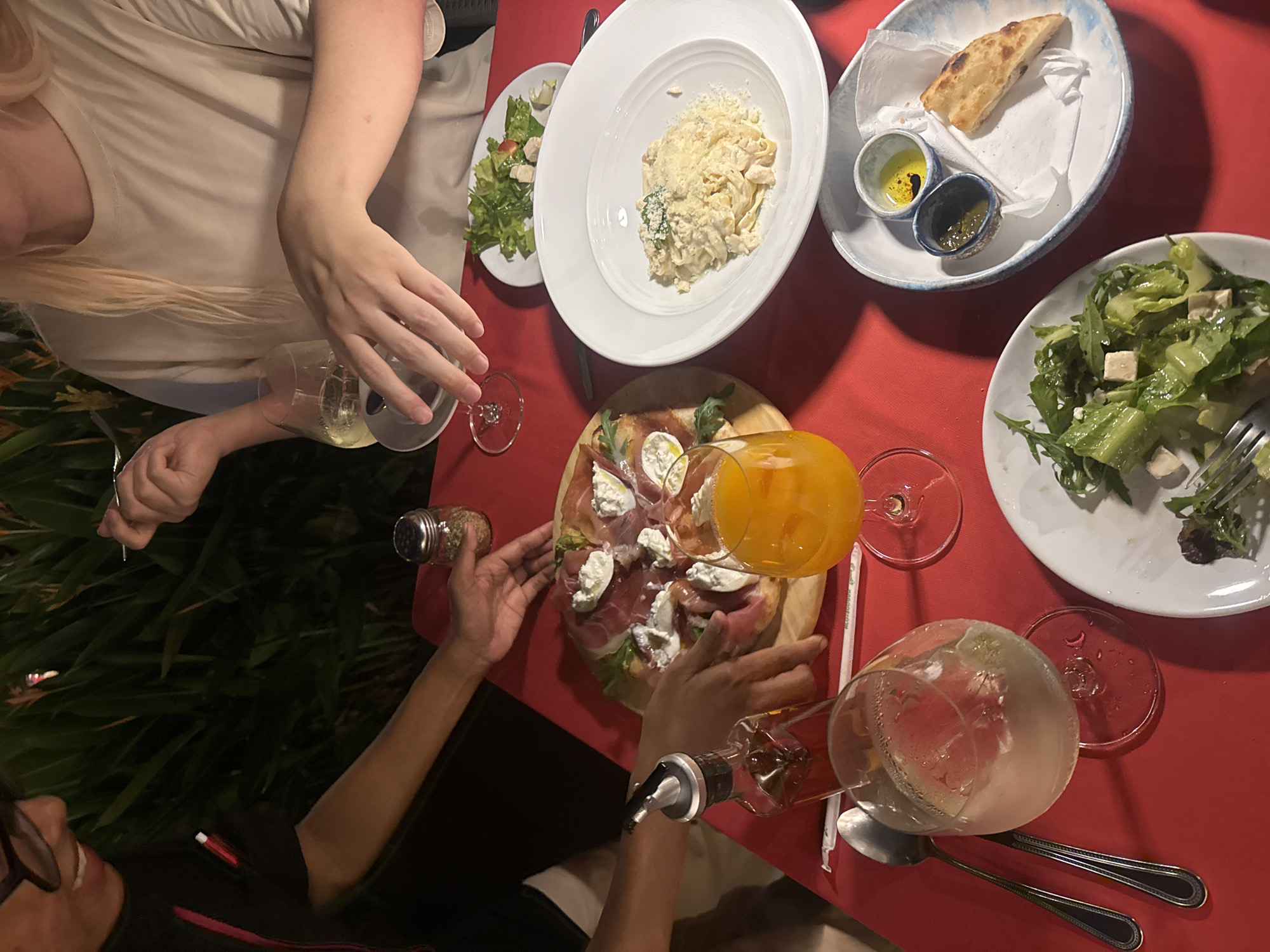The image depicts a group of people enjoying a meal at a restaurant. A red tablecloth covers the table, suggesting an outdoor setting or a restaurant with a vibrant decor. The lighting suggests it might be evening or indoors with warm lighting. On the table, several dishes are visible, including a pasta dish, a salad, a pizza, and bread with dips. Drinks are also present, with glasses of wine and juice. The people are engaged in the meal, with hands reaching for food and drinks. One person is seen pouring a drink, while another is holding a spice shaker. The presence of both Western and Italian dishes indicates a diverse menu, common in many tourist destinations like Chiang Mai. The overall mood appears relaxed and convivial, with people sharing a meal together. The background shows glimpses of greenery, possibly from plants or trees outside the restaurant. There is no visible text in the image.