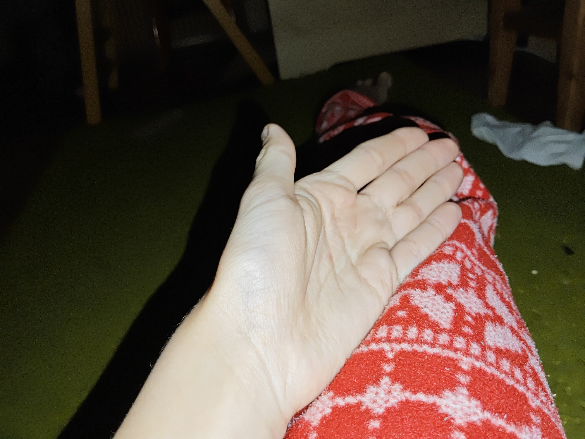 A person's hand is resting on a red and white patterned cloth, likely a towel. The scene is dimly lit, with a green floor visible. There are wooden furniture legs in the background. Another foot is also visible in the background. The location is most likely indoors. There is no indication of it being Odesa, Ukraine.