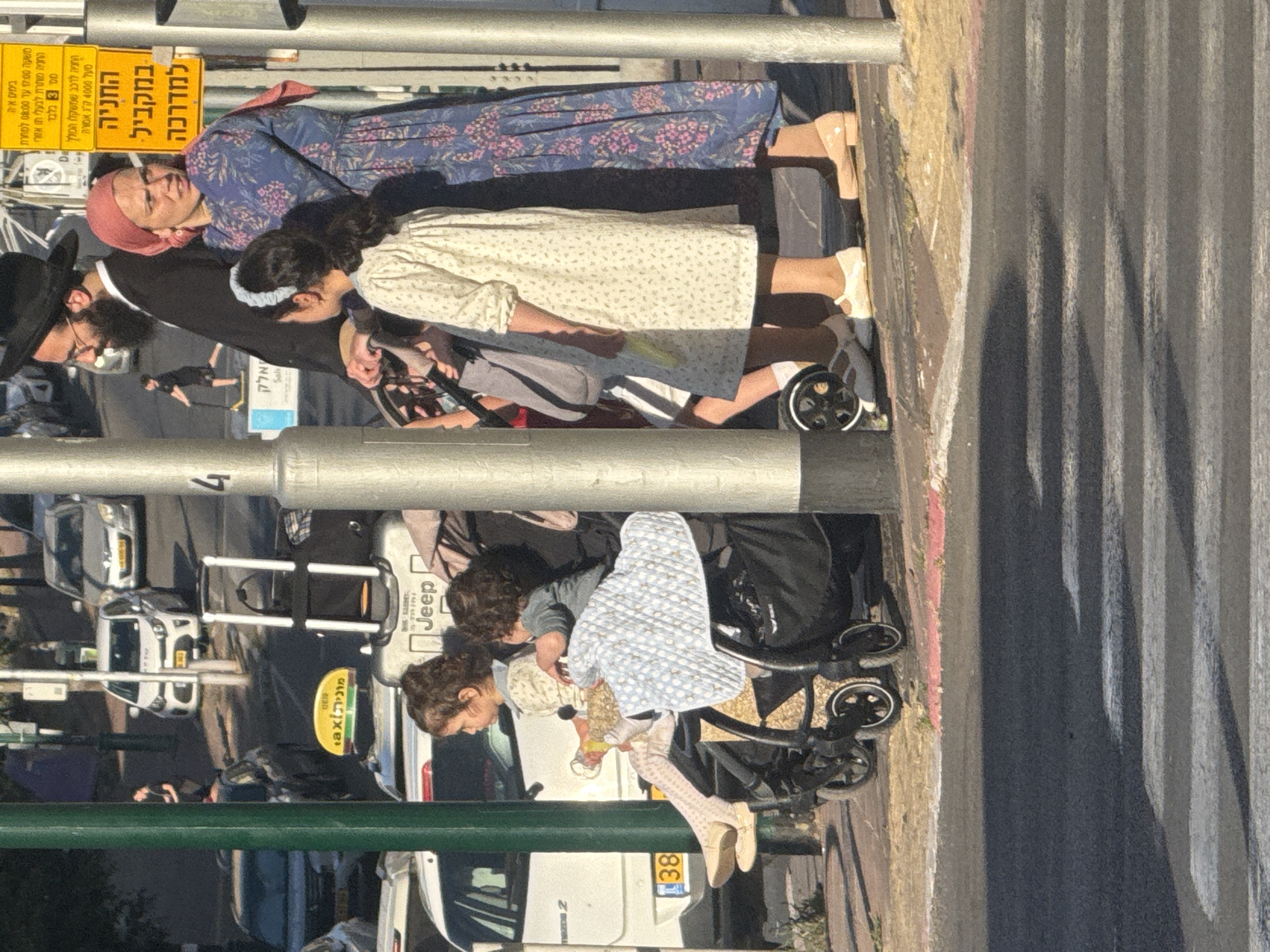 This image shows a group of people walking on a street in Ramat Gan, Israel.
From top to bottom, the image depicts a crosswalk, a family of four including: a mother and daughter wearing traditional Jewish clothing walking ahead, with the father and a child seated in a stroller following them. Street signs written in Hebrew are visible on the top left. There is also a street scene with cars and taxis parked nearby.