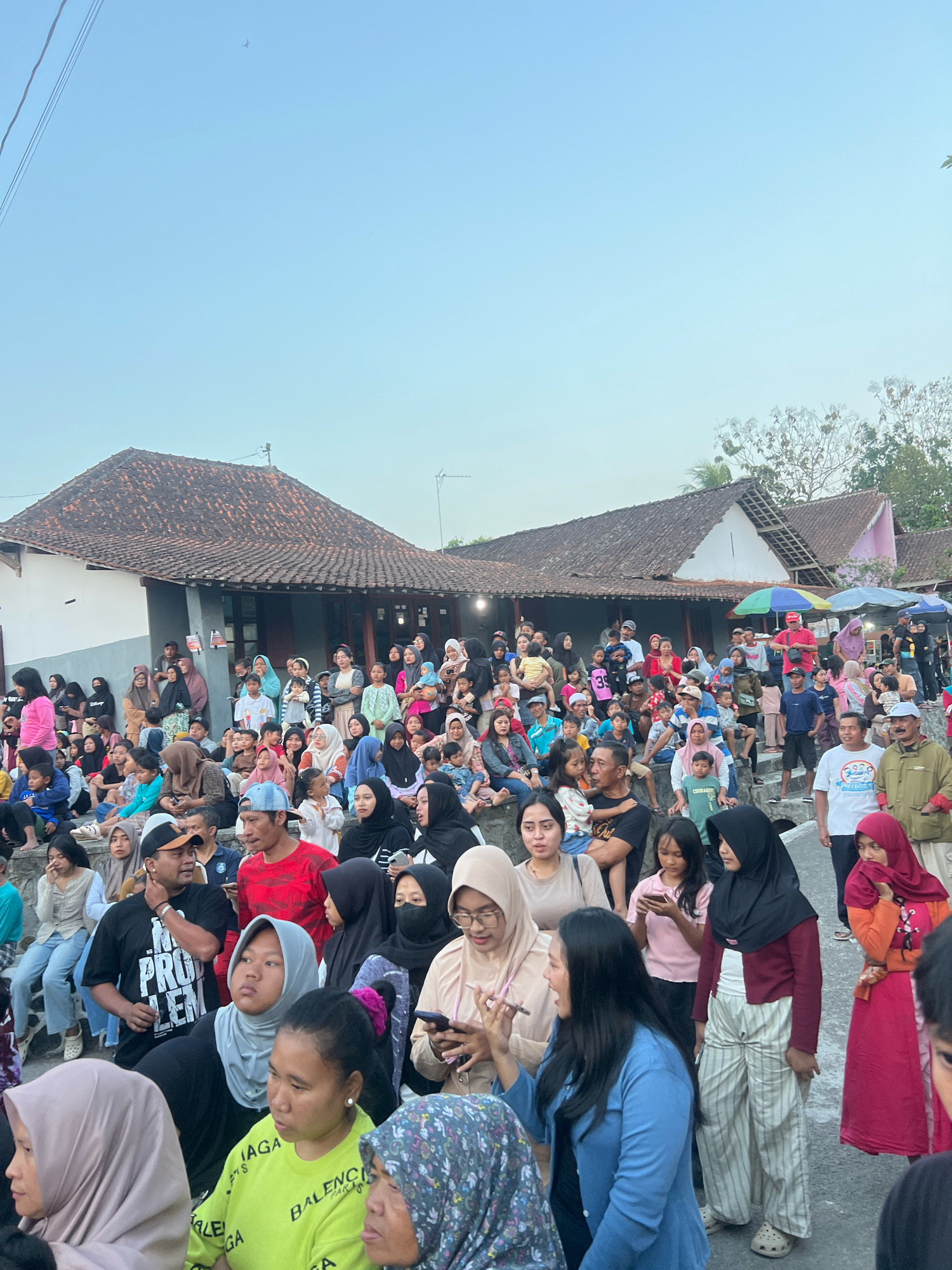 The image depicts a gathering of people in an outdoor setting, likely in the city of Madyocondro, Indonesia, as suggested by the context. A crowd is seated and standing, some on steps and others on the ground, facing towards the right of the frame. They appear to be observing an event or performance, with some individuals holding up their phones.
The environment features traditional Indonesian architecture, with buildings characterized by terracotta tiled roofs and simple facades. Trees are visible in the background, suggesting a semi-rural or suburban area. The sky is clear and blue, indicating daytime.
The individuals in the crowd are diverse in age and gender. Many women are wearing hijabs, common in Indonesia. Clothing styles range from casual t-shirts and pants to more traditional attire. Some people are interacting with each other, pointing or talking, while others are focused on something outside the frame. Children are also present, some being held by adults and others standing or sitting independently.
Several objects are scattered throughout the scene. Umbrellas are present, some open and providing shade, while others are closed. Food stalls or vendors might be present on the right side of the frame, indicated by colorful umbrellas and some visible items on tables. Power lines are visible in the upper left portion of the image, and a small bird or insect is in flight against the blue sky. The overall atmosphere appears to be one of a community event or celebration.