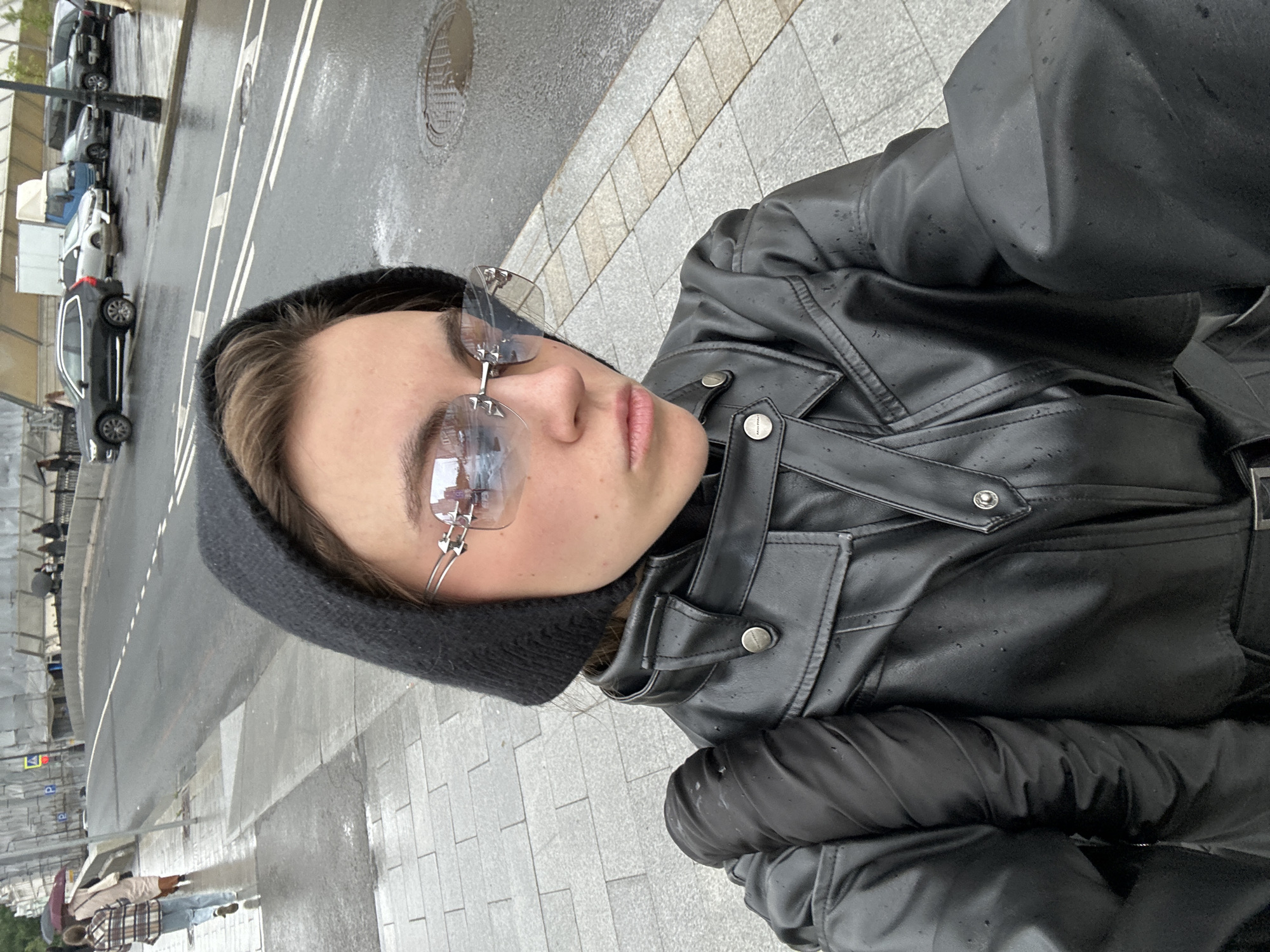 A woman in a black leather jacket and black beanie stands on a wet city sidewalk, facing forward and taking a selfie. She wears distinctive silver-rimmed, blue-tinted sunglasses. Her left arm is covered by a long black glove. The ground is paved with grey interlocking tiles. The sidewalk is wet, with puddles reflecting the overcast sky. Behind her, a street runs parallel to the sidewalk, with cars driving on it and parked along the side. The street is also wet, with white dashed lines marking lanes. In the background, there is a bridge under construction, covered in scaffolding. Several people are visible in the background walking on the sidewalk and crossing the street. The weather appears to be rainy or recently rained. The scene suggests an urban environment, likely in Moscow, Russia, given the architecture and road markings.