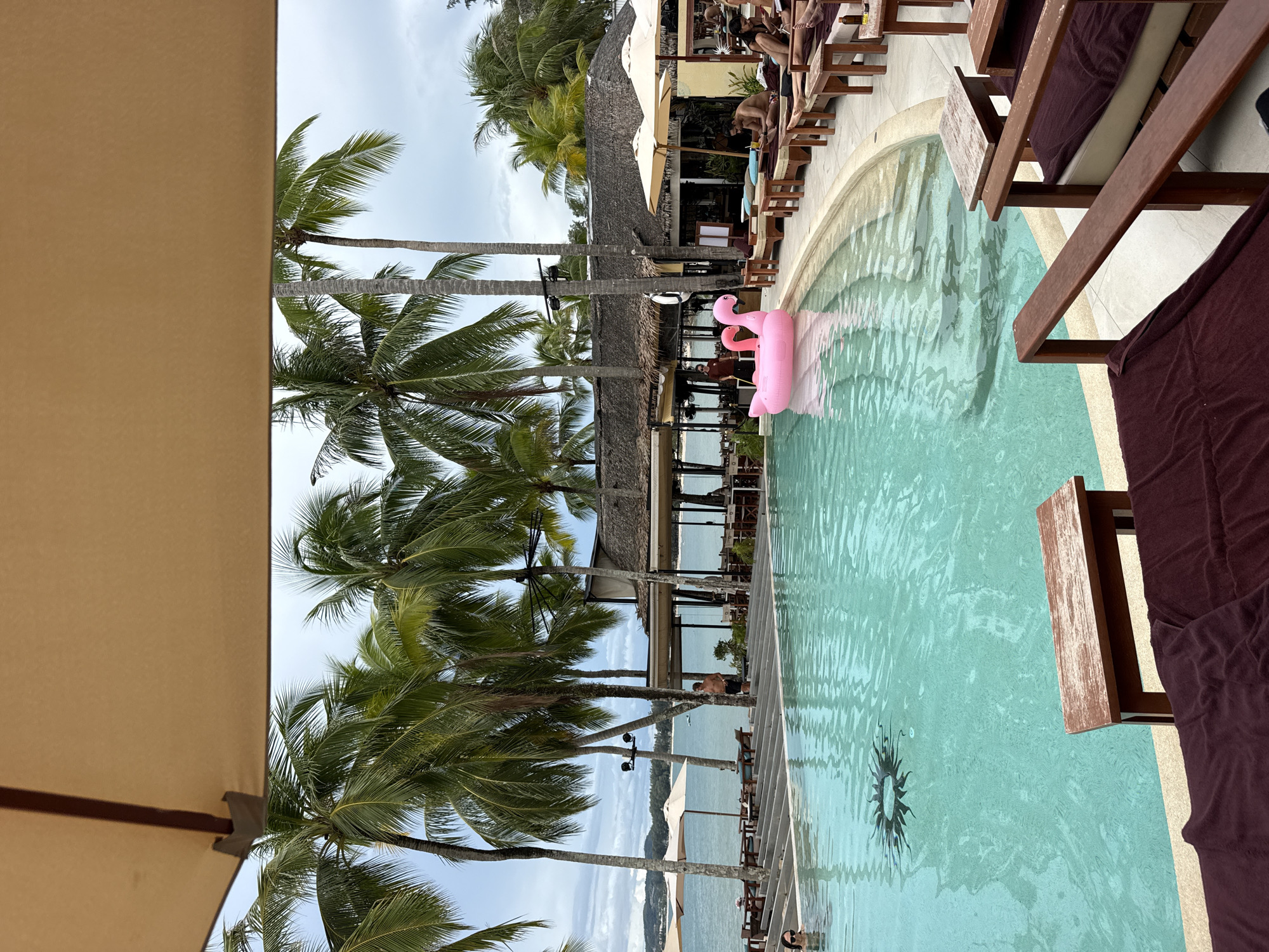 The image depicts a resort scene in Choeng Thale, Thailand. A large, clear turquoise swimming pool with a decorative sunburst mosaic at the bottom occupies the foreground. A pink inflatable flamingo float is visible in the pool. To the right of the pool, several lounge chairs with dark red cushions and small wooden tables are arranged on a tiled patio. In the background, a row of palm trees frames a view of the ocean and a beachfront restaurant with thatched roofs. Several people are seen relaxing on lounge chairs, while others are partially submerged in the pool or standing near the restaurant. The sky is overcast, suggesting a cloudy day.