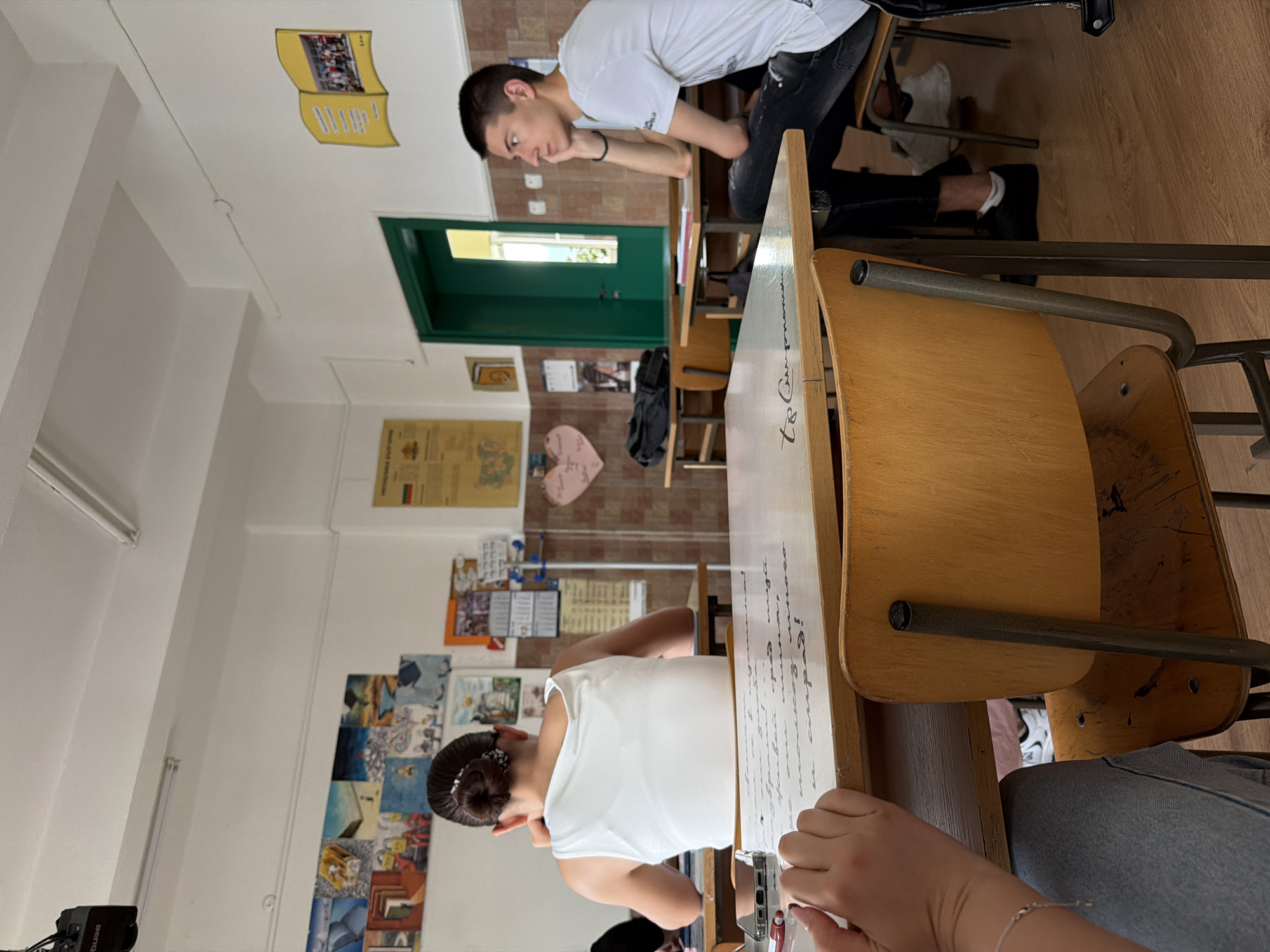 The image depicts a classroom interior, likely at a school in Panagyurishte, Bulgaria, given the context provided. The scene is viewed from a slightly elevated angle, focusing on two students at their desks.
In the foreground, a young person wearing a white tank top and with their hair in a bun is seated at a desk, facing away from the camera. Their left arm is visible, resting on the desk. The desk surface in front of them has writing on it. A portion of another person's arm and hand, wearing blue denim jeans, is visible to the left, also resting on the desk.
To the right, a young male student in a white t-shirt and dark jeans is seated at a desk, facing towards the right of the frame. He is leaning his head on his hand, looking away from the camera. His legs are crossed, and he is wearing black sneakers with white socks.
The classroom environment includes wooden desks and chairs, typical of older school furniture. The walls are adorned with various educational materials, including a map, artwork, a calendar, and a drawing of an open book. A green-painted door is visible in the background. The lighting suggests it is daytime, with light fixtures on the ceiling. The overall atmosphere appears to be one of quiet study or a lesson in progress.