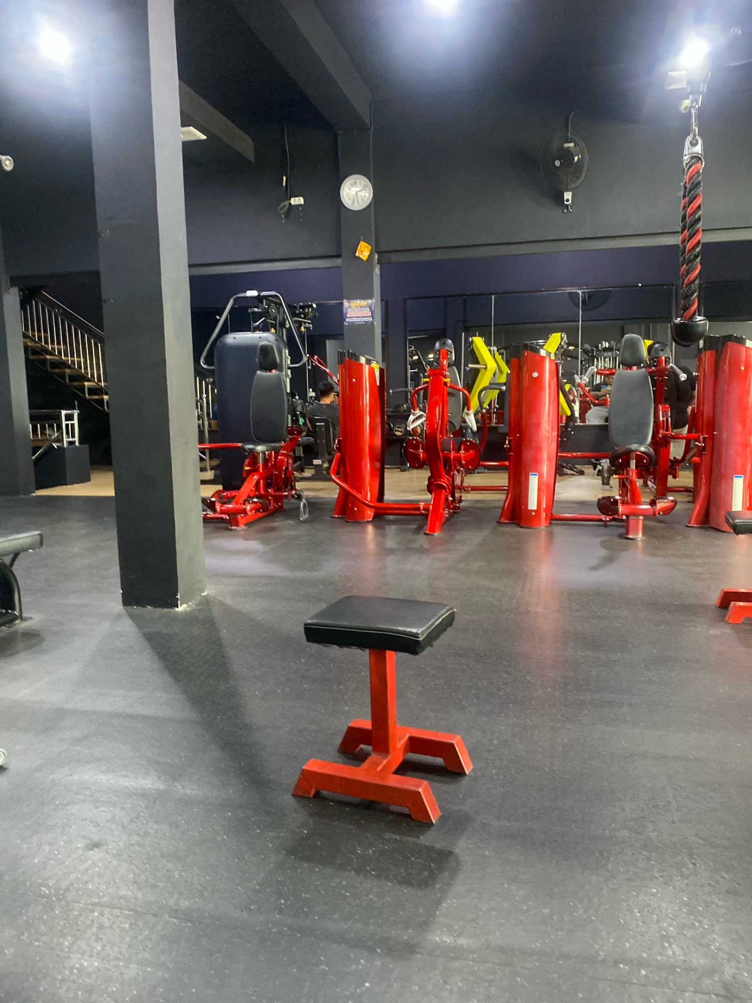 The image depicts the interior of a gym, specifically located in Banjarbaru, Indonesia. The gym features a dark gray rubberized floor and black walls, with exposed metal beams and ductwork visible on the ceiling. Bright, white fluorescent lights illuminate the space.
Prominently featured are numerous red and yellow weight training machines, arranged in rows. These machines appear to be a mix of resistance machines with padded seats and backrests, as well as some cable-based equipment. In the foreground, a small, red exercise stool with a black padded seat is positioned centrally.
Several individuals are present in the gym, though their interactions are not clearly defined. One person is seen sitting on a machine in the background, facing away from the camera. Another person is partially visible, also seated on a machine. A spiral staircase with metal railings is visible on the left side of the image, leading up to a dark platform.
The overall environment suggests a functional fitness facility.