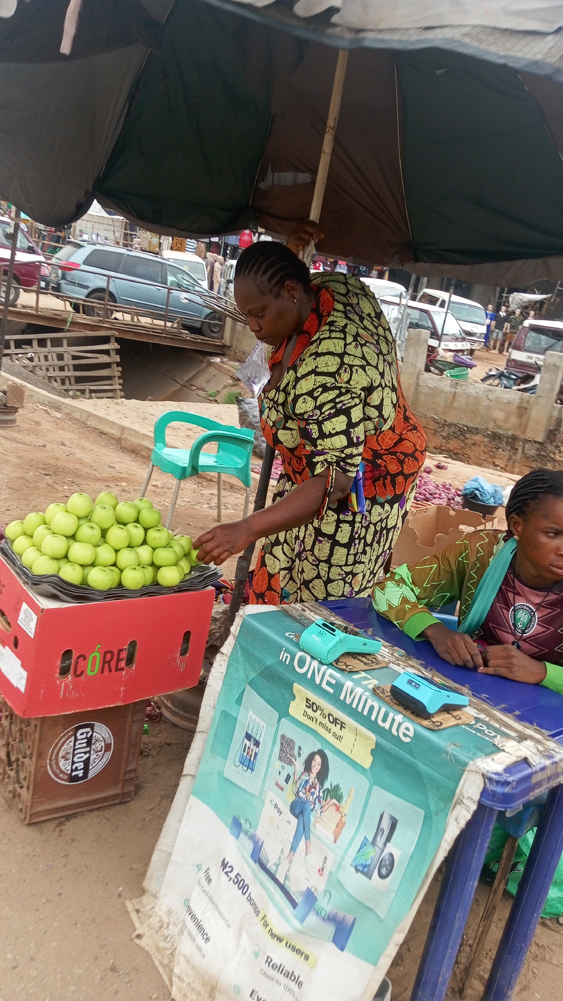 This image depicts a vibrant outdoor market scene in Onitsha, Nigeria, featuring two women and their stall. The setting is likely a busy street or open-air market, characterized by a dirt ground and vehicles visible in the background, including a light blue station wagon and other cars.
The main subject is a standing woman, presumably a vendor, with her hand near a large pile of neatly arranged green apples. She wears a brightly patterned dress with colors including green, black, and orange. Her hair is braided in cornrows. A large, dark green and brown umbrella provides shade over her and the stall.
To the left of the standing woman, a red crate labeled "CORE" supports the tray of green apples. Below it, a brown crate bearing the "Gulder" beer logo is visible. A light green plastic chair stands empty behind the apples.
To the right, a second, younger woman is seated at a blue plastic table. She wears a green and black patterned top, which appears to be a football jersey with "FOOTBALL" partially visible. The table is covered with a promotional banner for "Opay," an electronic payment service. The banner prominently displays text such as "in ONE Minute," "50% OFF Don't miss out!", "Pay," "N2,500 bonus For new users," "Free Card Maintenance Fee," "Convenience," and "Reliable." Illustrations on the banner include a woman, gas pumps, shopping carts, and electronic devices. Two light blue POS (Point of Sale) machines are resting on the banner-covered table.
The scene appears to be taking place during the daytime, with the umbrella suggesting either protection from sun or potential rain. The women's expressions are neutral, focused on their tasks or observing the surroundings. The overall impression is one of typical daily commerce in a bustling market environment.