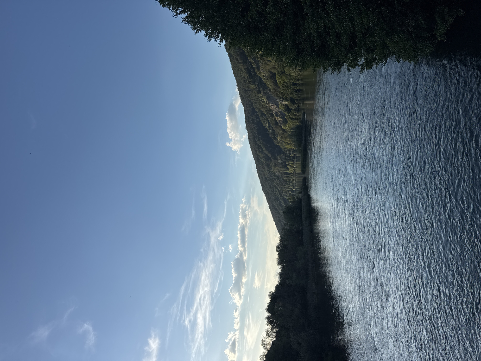 The image depicts a wide river flowing through a lush, forested landscape. The sky above is a clear, bright blue with a few wispy white clouds. On the left side of the river, the banks are lined with dense, dark green trees. The right side of the river is dominated by a steep, tree-covered hill that stretches into the distance. The river water is a deep blue, with visible ripples and small waves on its surface, indicating gentle movement. The overall impression is one of a serene and natural environment. The specific location in the city of Kokalyane, Bulgaria, is likely a scenic spot along the Iskar River, known for its natural beauty. There are no people or discernible human activity visible in the image. The scene is a tranquil depiction of nature.