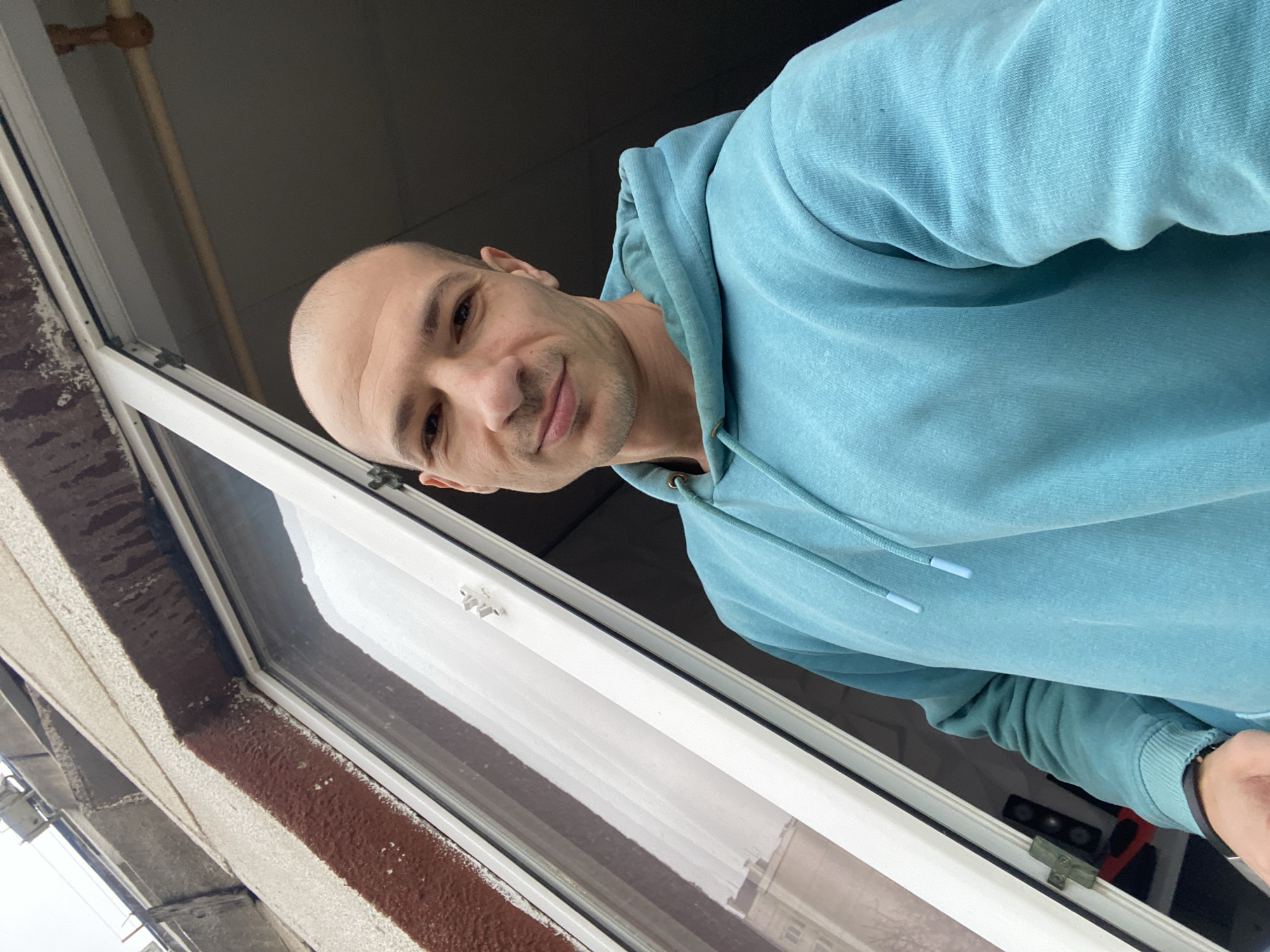 A bald man in a light blue hoodie is positioned at an open window. The window frame is white and the outside exterior shows a brown textured surface. Reflected in the window, part of an interior space is visible. The location is presumed to be Pleven, Bulgaria, based on the request.