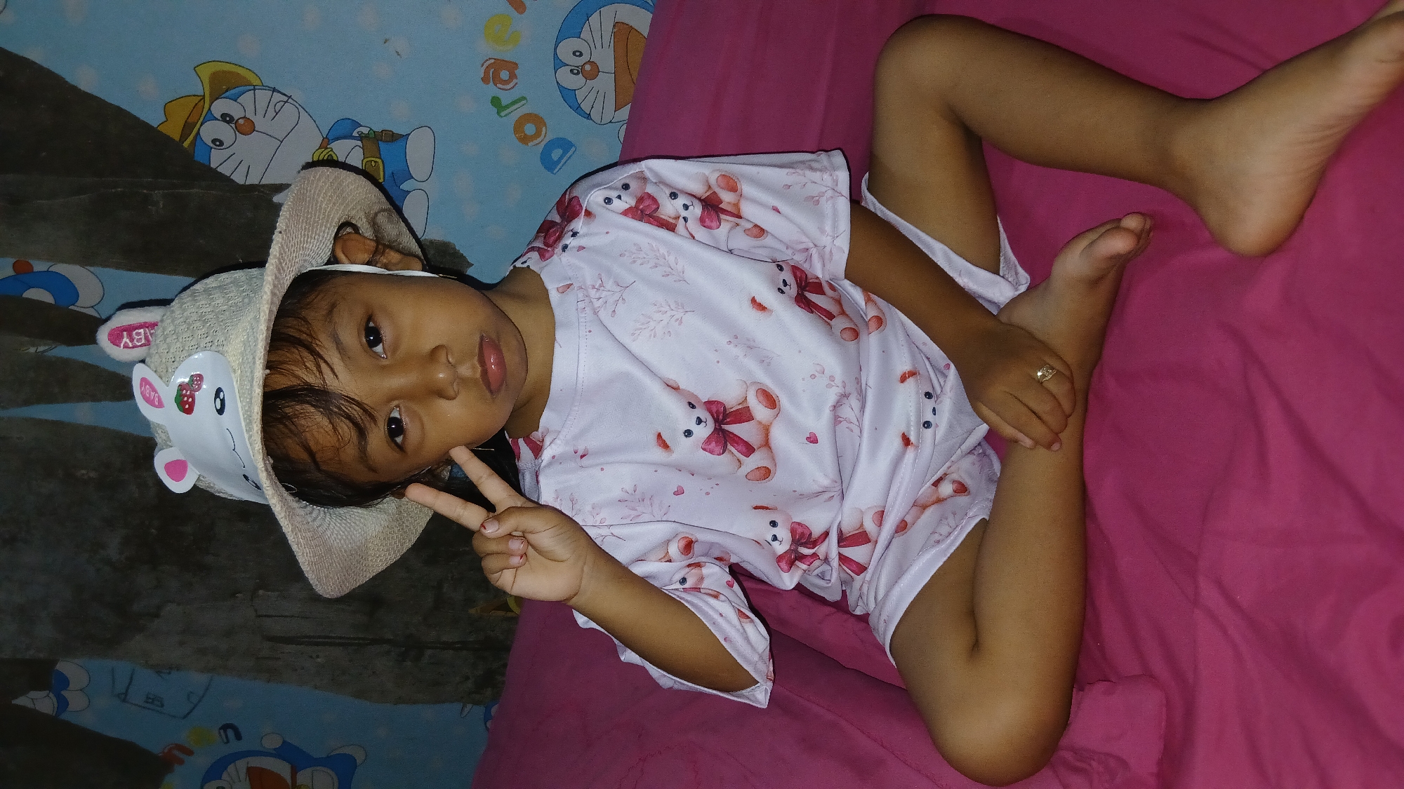 A young child, possibly a girl, is lying on a pink surface, which appears to be a bed or a blanket. The child is wearing a straw hat with a white band that has a rabbit-like design and the word "BABY" printed on it. The child is also wearing a light pink t-shirt or pajama top adorned with a pattern of teddy bears wearing red bows and delicate leaf designs. The child's left hand is raised, forming a peace sign with their index and middle fingers. The child's right hand is resting on their left leg, and a ring with a heart-shaped stone is visible on one of the child's fingers. The child's legs are bent, with their feet positioned upwards.
In the background, a blue patterned wallpaper is visible, featuring cartoon characters, one of which resembles Doraemon. The overall impression is that of a relaxed, indoor setting, likely a bedroom, during what appears to be nighttime or a dimly lit room, given the lighting. The child's facial expression is calm and perhaps a little sleepy. The setting of Keudejungkagajah, Indonesia, provides geographical context, but no specific visual cues within the image directly indicate this location.