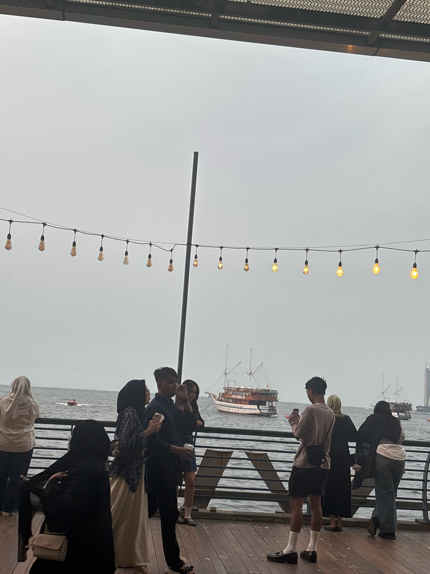 In North Jakarta, Indonesia, a group of people stand on a wooden boardwalk overlooking the sea. The sky is overcast, and several boats, including a large two-masted vessel, are visible on the water. String lights with exposed bulbs hang across the scene, casting a warm glow.
In the foreground, several individuals are interacting. A woman in a cream-colored hijab stands with her back to the camera, facing the water. To her left, another woman in a black hijab is seated on a bench, with a beige crossbody bag on her lap. In the center, a group of three individuals are standing. One man, wearing a dark blue shirt, is holding a white cup. Next to him, a woman in a patterned blue and white hijab holds a phone. To their right, another man in a grey long-sleeved shirt and black shorts is holding a phone and looking towards the left. He is wearing white socks and black loafers. A woman in a green hijab stands to his right, leaning on a railing. Further to the right, a woman in a black hijab and jeans is also leaning on the railing, looking towards the sea.
The boardwalk has a dark wooden surface and a metal railing along the edge of the water. In the distance, more boats and a tall building are visible. A small red boat is in the water on the left side of the frame. The overall atmosphere appears to be a casual outing, possibly a gathering or a stroll along the waterfront.