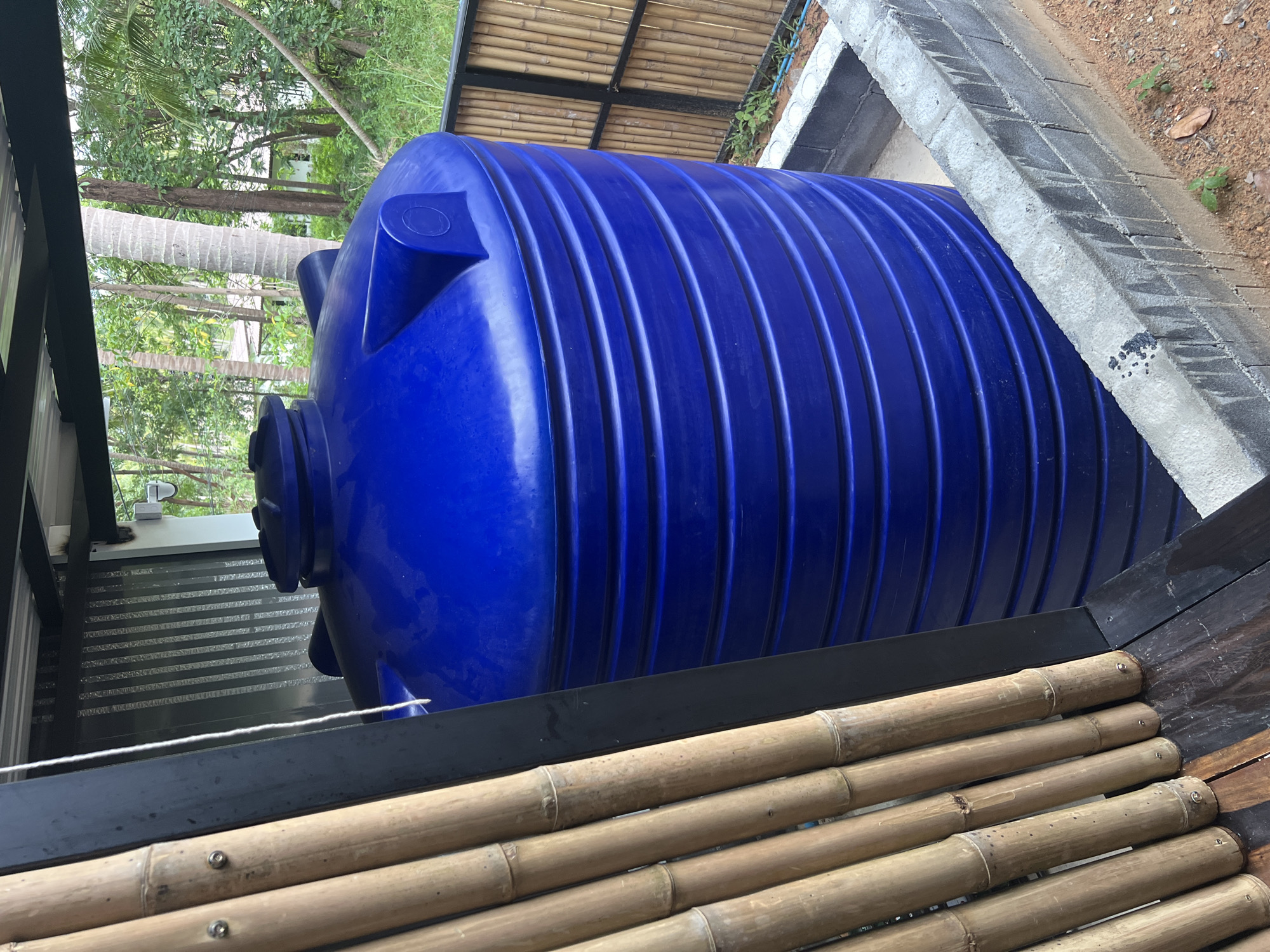 A large, blue plastic water tank stands on a concrete base, partially surrounded by a low brick wall. The tank has a ribbed exterior and a domed top with a lid. To the left of the tank is a vertical structure made of bamboo poles. In the background, there is a dense tropical forest with palm trees and other vegetation, and a glimpse of white buildings. The scene appears to be outdoors, possibly in a residential area or a resort, given the tropical setting and the presence of a water storage solution. The location is identified as Ko Pha-ngan District, Thailand.