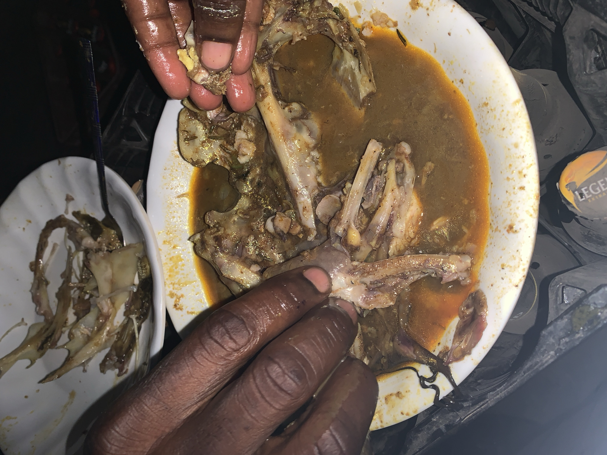 In a location identified as New Stadium, Nigeria, the image depicts two white ceramic plates containing fish remnants. The larger plate contains fish bones and pieces submerged in a brown, oily broth. A dark-skinned person's hand is present, appearing to be picking apart the fish remains. The smaller plate holds a collection of picked-over fish bones. A metal utensil is placed alongside the smaller plate. An unopened beverage can sits near the edge of the larger plate, with a portion of its label visible. The entire scene appears to be situated on a dark, possibly cluttered, surface.