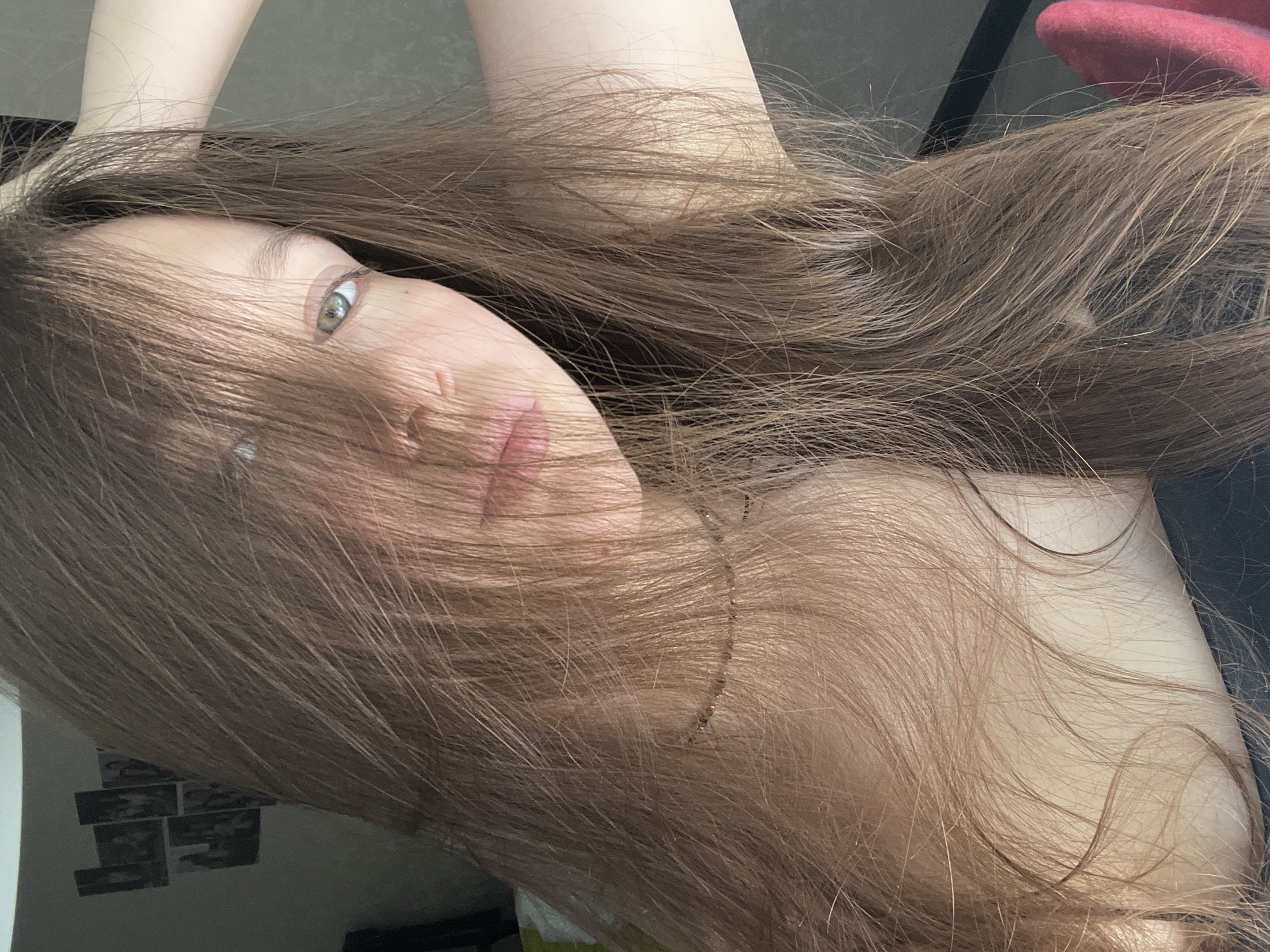 A young woman with long brown hair partially obscuring her face lies in a reclined position. Her face is angled, revealing one light-colored eye and a portion of her lips. Her hair is spread around her head and shoulders. A thin gold necklace is visible on her neck. In the background, a section of a wall is visible with several small, rectangular objects attached to it and a red chair can be seen.
There is no evidence this photo was taken in Chaikovsky, Russia.