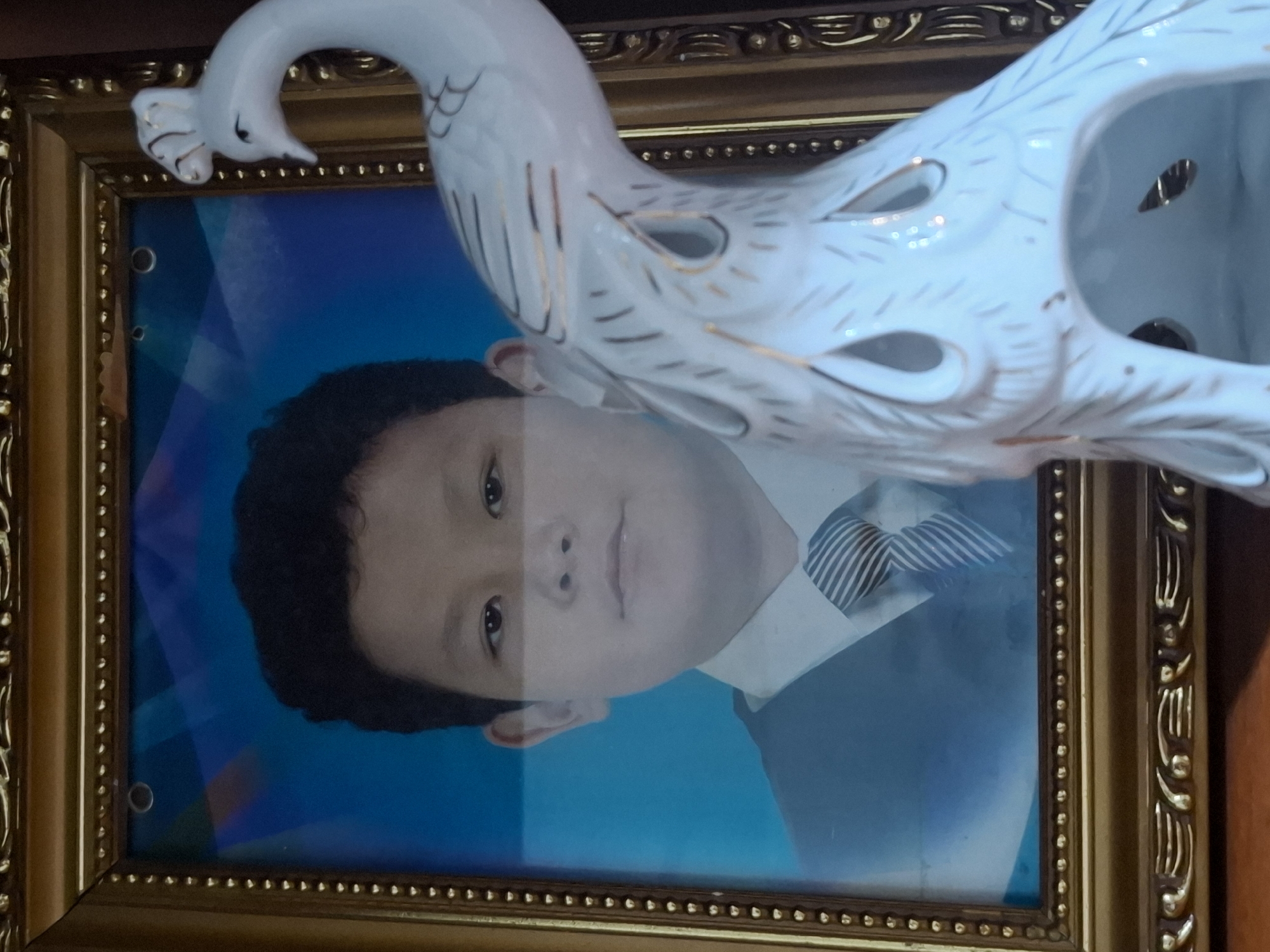 A framed portrait of a young boy wearing a suit and tie is positioned on a surface, possibly a table or shelf. A white ceramic figurine of a peacock stands in front of the portrait, partially obscuring it on the right side. The boy's face is clearly visible, looking directly at the camera. The background of the portrait features a blue abstract design. The peacock figurine is detailed with gold accents. The overall scene suggests an indoor setting, likely a home, and there is no indication of Almaty, Kazakhstan.