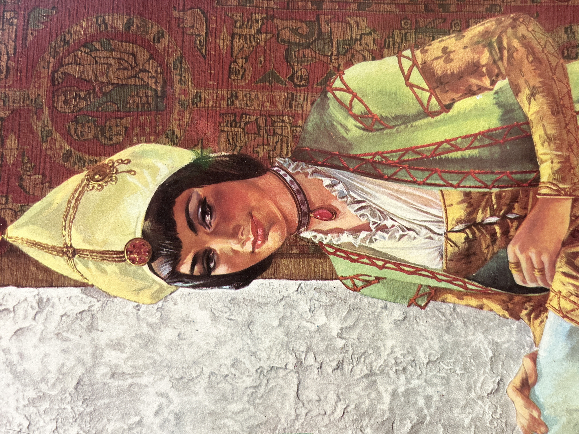 The image is a painting of a woman in traditional attire, possibly of Middle Eastern or Central Asian origin. She wears a pale yellow, conical hat adorned with gold trim and a circular, red gem. Her dark hair is short and frames her face, which has prominent makeup. She is also wearing a green tunic with red trim over a white, ruffled blouse and a golden vest. A dark choker with a red pendant necklace completes her ensemble. The background to her right features a tapestry with intricate patterns, primarily in red and green hues, depicting figures. To her left is a rough, textured white wall. The woman's hands are visible, one resting on a light blue cloth, the other adorned with rings and holding another object. There is no discernible reference to Santa Ana, Venezuela in the image.