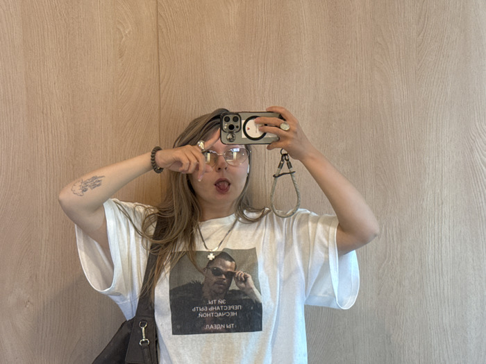 A woman with long brown hair poses for a selfie in front of a light brown wood panel wall. She wears glasses, a white t-shirt with a graphic, a silver necklace, and a bracelet. She holds her phone in both hands, with a ring on her right hand. The phone has a wrist strap. She sticks out her tongue and makes a peace sign near her face. A tattoo is visible on her left arm. A dark bag hangs from her shoulder.