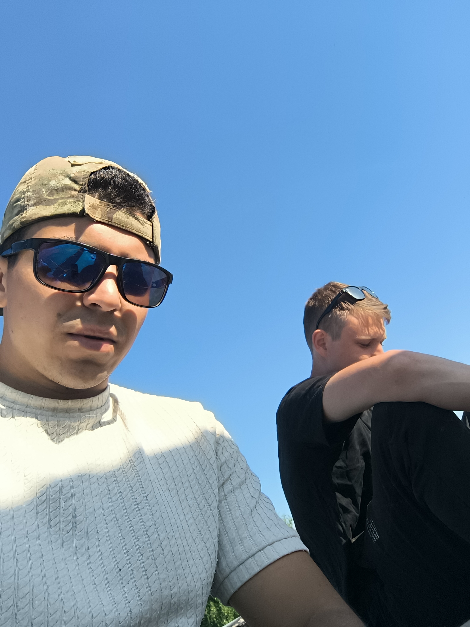 Two young men are visible in the image against a clear, bright blue sky. The man on the left is wearing a white textured short-sleeved shirt, sunglasses with a reflective blue tint, and a camouflage baseball cap. He appears closer to the camera, looking slightly downward. The man on the right, positioned further back, is wearing a black shirt and pants. He has sunglasses on top of his head and his face is obscured, as he seems to be resting his head on his arm. The location appears to be outdoors, based on the lighting and clear sky. The specific city is unknown.