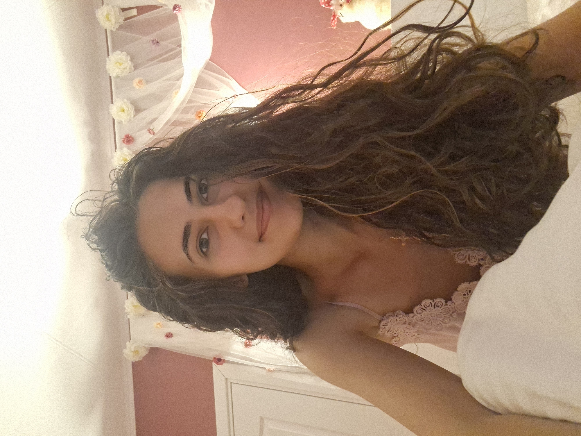 The image shows a young woman with long, wavy brown hair, wearing a pink camisole with lace detailing. She is lying in a bed, with a white pillow in the foreground. The background appears to be a bedroom with pink walls and a white canopy adorned with flowers. A closed white door is visible on the left side of the image. The lighting is soft and warm, suggesting an indoor setting. The woman is looking directly at the camera with a slight smile.