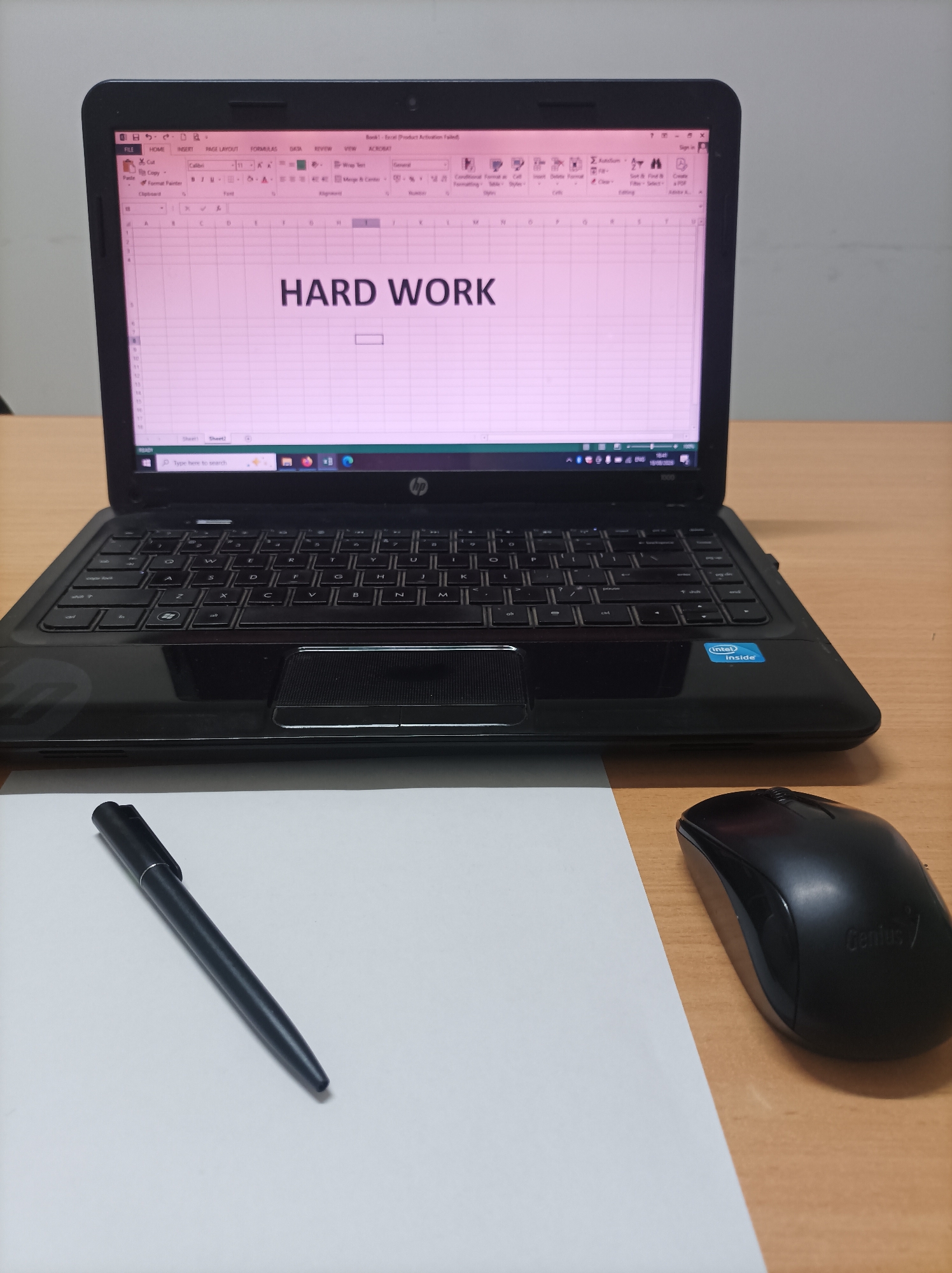 The image depicts a close-up, eye-level view of a desk setup, likely in an office or study. In the foreground, a black pen rests diagonally on a white sheet of paper. To the right of the paper, a black computer mouse with the "Genius" logo is positioned on a light brown wooden desk.
Dominating the mid-ground is an open black laptop, its screen displaying an Excel spreadsheet. The title "HARD WORK" is prominently centered on the spreadsheet in a large, bold font. The laptop's keyboard and trackpad are visible. A sticker that reads "Intel Inside" is adhered to the laptop's casing below the keyboard.
The background is indistinct, suggesting a plain wall. The overall lighting is soft, illuminating the workspace.
There is no information available to determine the specific location in Nganjuk, Indonesia.