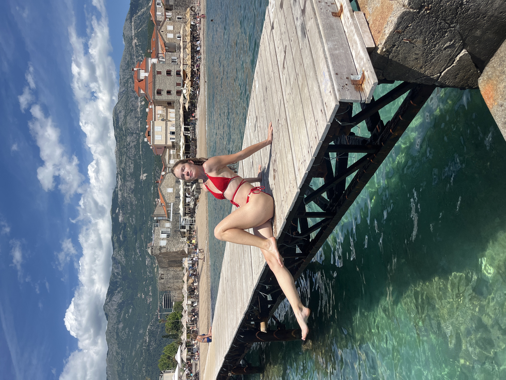 A woman in a red bikini is sitting on a wooden pier, with her legs dangling over the clear, turquoise water. The woman has light-colored hair and is looking towards the camera. In the background, there is a picturesque coastal town with buildings featuring red-tiled roofs, nestled at the base of a large, green mountain range. The sky is bright blue with scattered white clouds, indicating a sunny day. Below the pier, the water is transparent enough to see the structures supporting the pier and the rocky seabed. On the shore behind the pier, there's a sandy beach with several umbrellas and what appear to be people relaxing. The overall scene suggests a vibrant summer day at a popular vacation destination. The image is taken during the daytime, under bright sunlight. There is no visible text in the image.