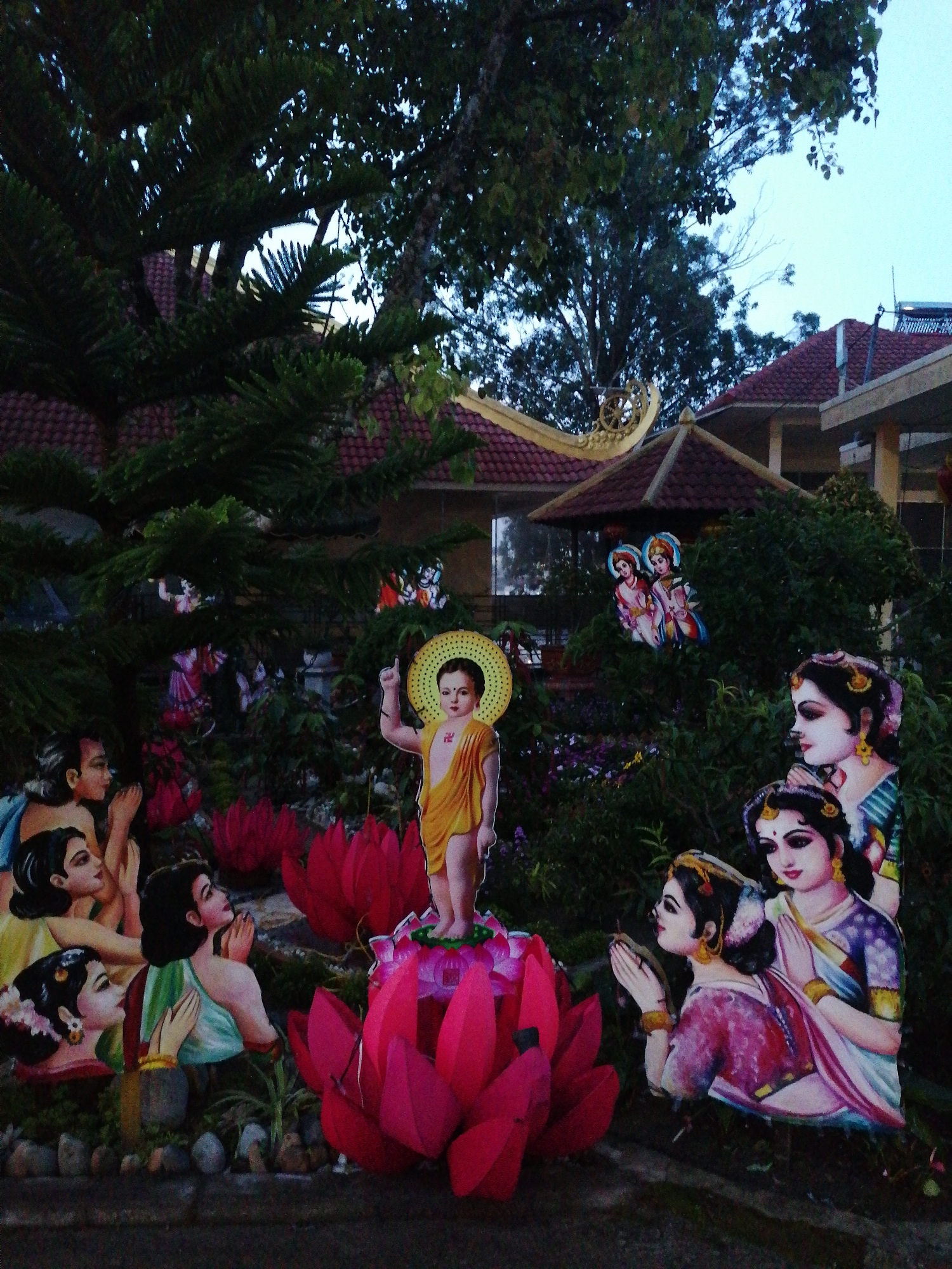 The image shows a roadside religious display in Da Lat, Vietnam, likely part of a Buddhist temple or garden. A cardboard cutout of the infant Buddha is prominent, standing atop a pink lotus flower. Several cardboard figures of deities and people are arranged around the Buddha, amidst shrubbery, trees, and small rocks. The scene is in front of a building with a red-tiled roof, possibly a temple structure. The image appears to have been taken in the late afternoon or evening, given the low light levels.