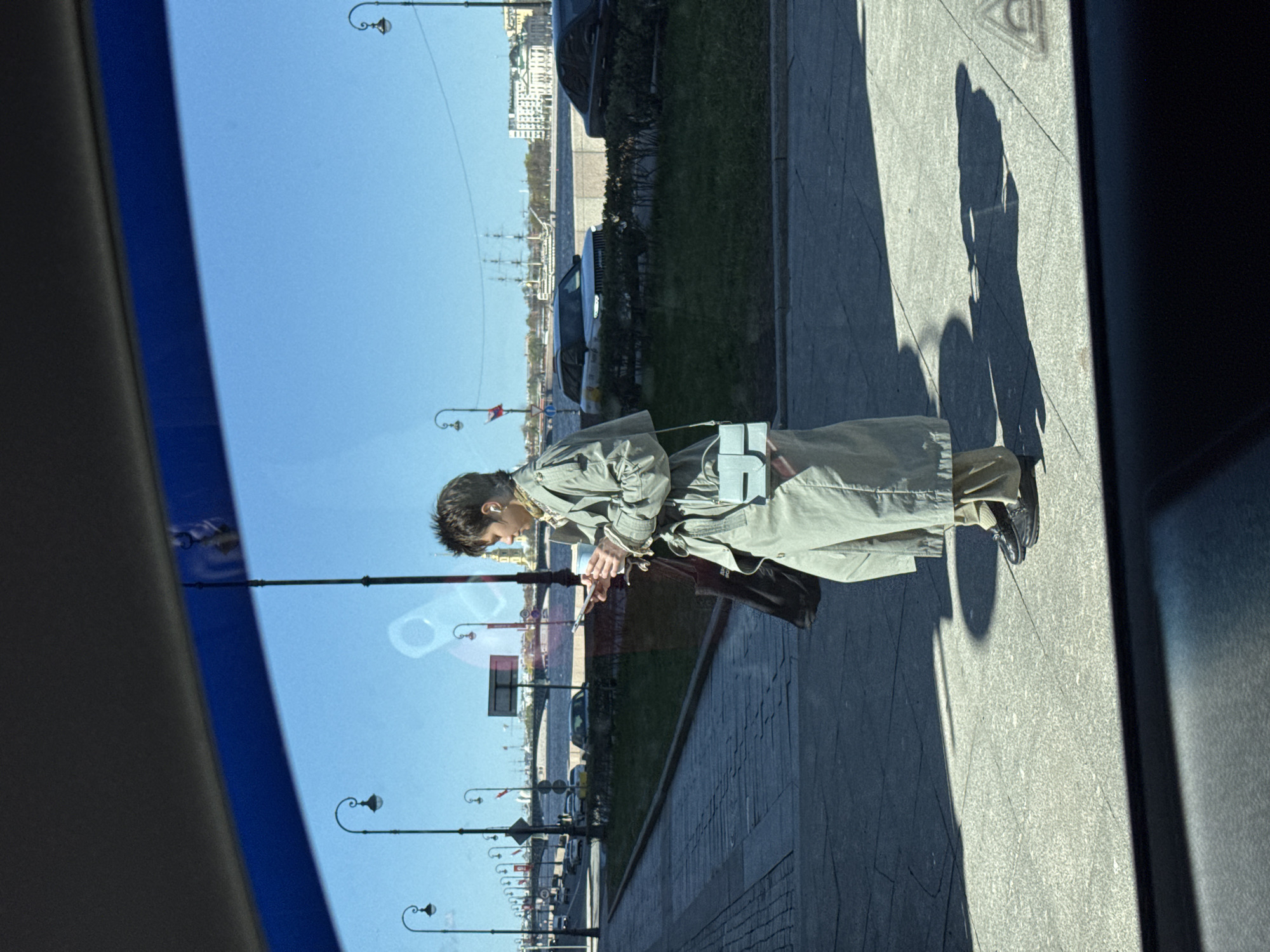 The image, a vertical shot taken from within a vehicle, depicts a man standing on a paved sidewalk adjacent to a body of water, likely a river or canal, in Saint Petersburg, Russia. The view through the window is partially obstructed by elements of the vehicle's interior, creating a framed perspective.
The man, facing the water, is dressed in a light-colored trench coat and dark shoes. He appears to be looking at or interacting with a mobile device or a small object he holds in his hands.
In the background, across the water, a cityscape is visible with buildings and structures characteristic of Saint Petersburg's architecture. Lamp posts line the sidewalk on which the man stands, and the scene is bathed in daylight. The image captures the reflection of the cityscape and the lamp posts in the vehicle's window, adding a layer of visual complexity. The man's shadow stretches out onto the sidewalk, indicating the position of the sun.