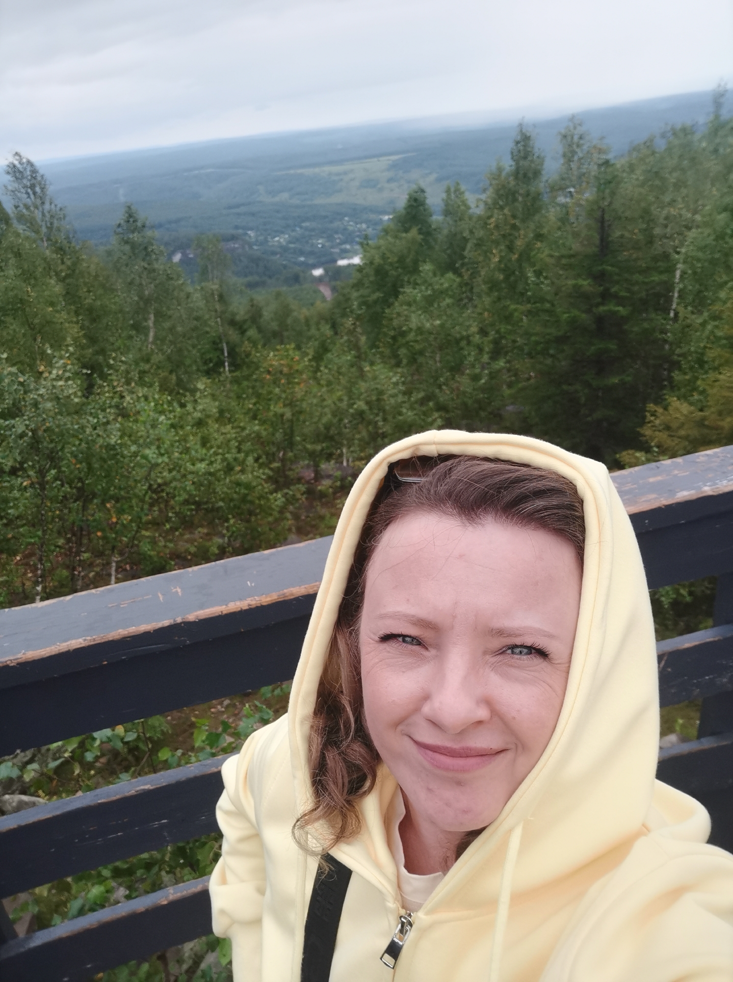 A woman in a yellow hooded sweatshirt takes a selfie from a wooden platform overlooking a vast, green, forested landscape. The sky is overcast. The woman has light skin, blue eyes, and curly brown hair that is partially visible under her hood. Her mouth is closed in a slight smile. In the background, the dense forest extends to the horizon, with a small town or village visible in the distance. The platform has a dark, wooden railing. The image appears to be taken in Gubakha, Russia, possibly from a viewpoint in the Kizel coal basin area known for its forests and natural beauty.