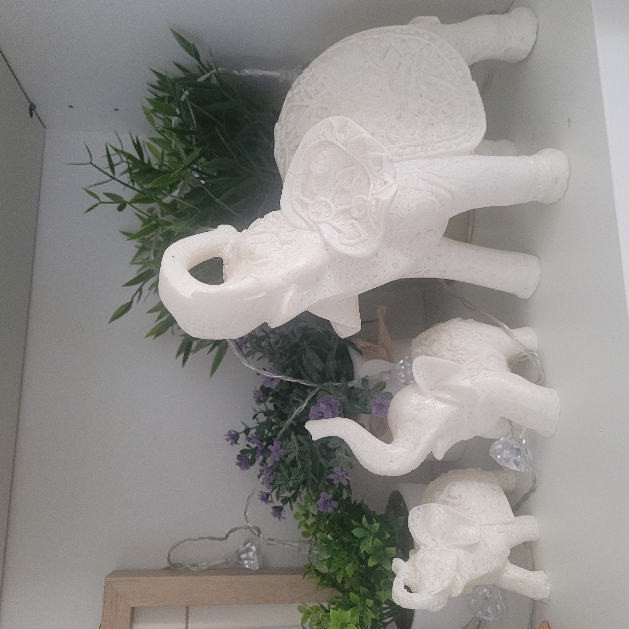 The image depicts a shelf displaying three white elephant figurines, a small potted plant with green foliage, another potted plant with purple flowers, and a string of fairy lights with diamond-shaped lights. The figurines vary in size, with the largest on the right and two smaller ones to its left. The elephant on the far right is the most detailed, featuring intricate carvings on its back and blanket. The fairy lights are draped across the shelf, with some lights visible near the elephant figurines. The background is a plain white wall. The scene is an indoor setting, likely a home or retail environment, and is not specifically identifiable as being located in Jūrmala, Latvia, or any other specific city.