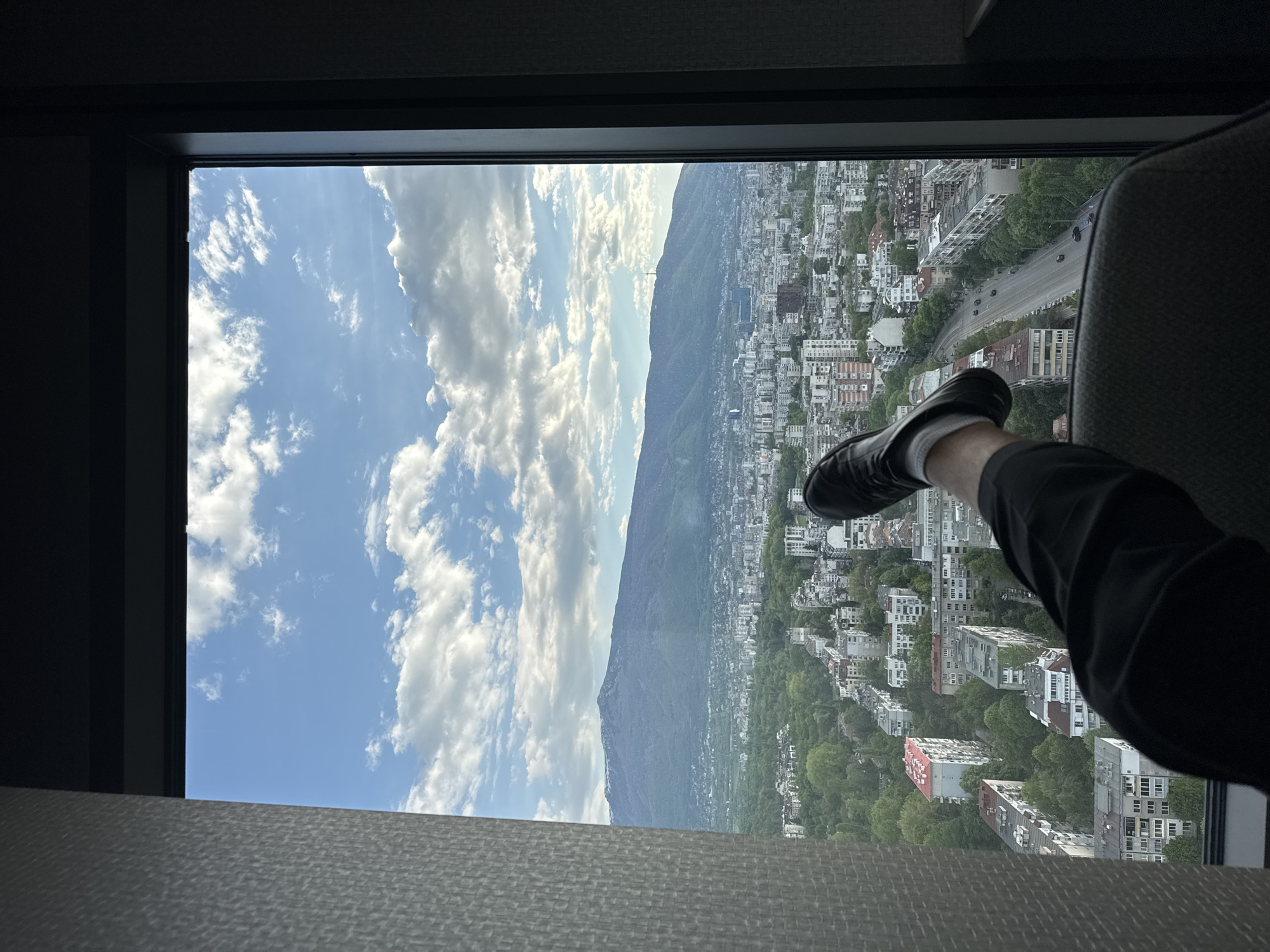 A view from a high vantage point, presumably a building in Sofia, Bulgaria. The image is oriented vertically, capturing a scene through a window.
The view is dominated by a city skyline with a prominent mountain range in the background, partially obscured by clouds. Buildings of various sizes and shapes fill the lower portion of the frame, interspersed with trees and patches of greenery. A major roadway is also visible, with cars moving along it.
The viewer's legs are positioned in the lower right corner of the image, suggesting a relaxed pose. The lower part of a chair is visible on the right side of the frame.