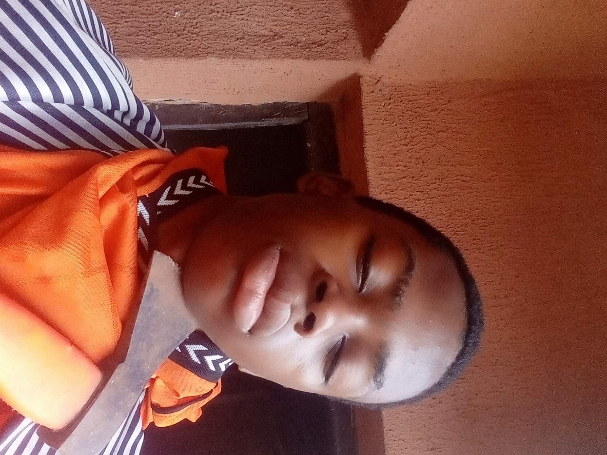 A person with dark skin lies with their eyes closed. They are wearing an orange cloth and a striped shirt. A metal object is held on top of the orange cloth. The background includes a window frame and a textured orange wall. The image was likely taken indoors. There is no direct evidence in the image to confirm it was taken in Nkpor, Nigeria.