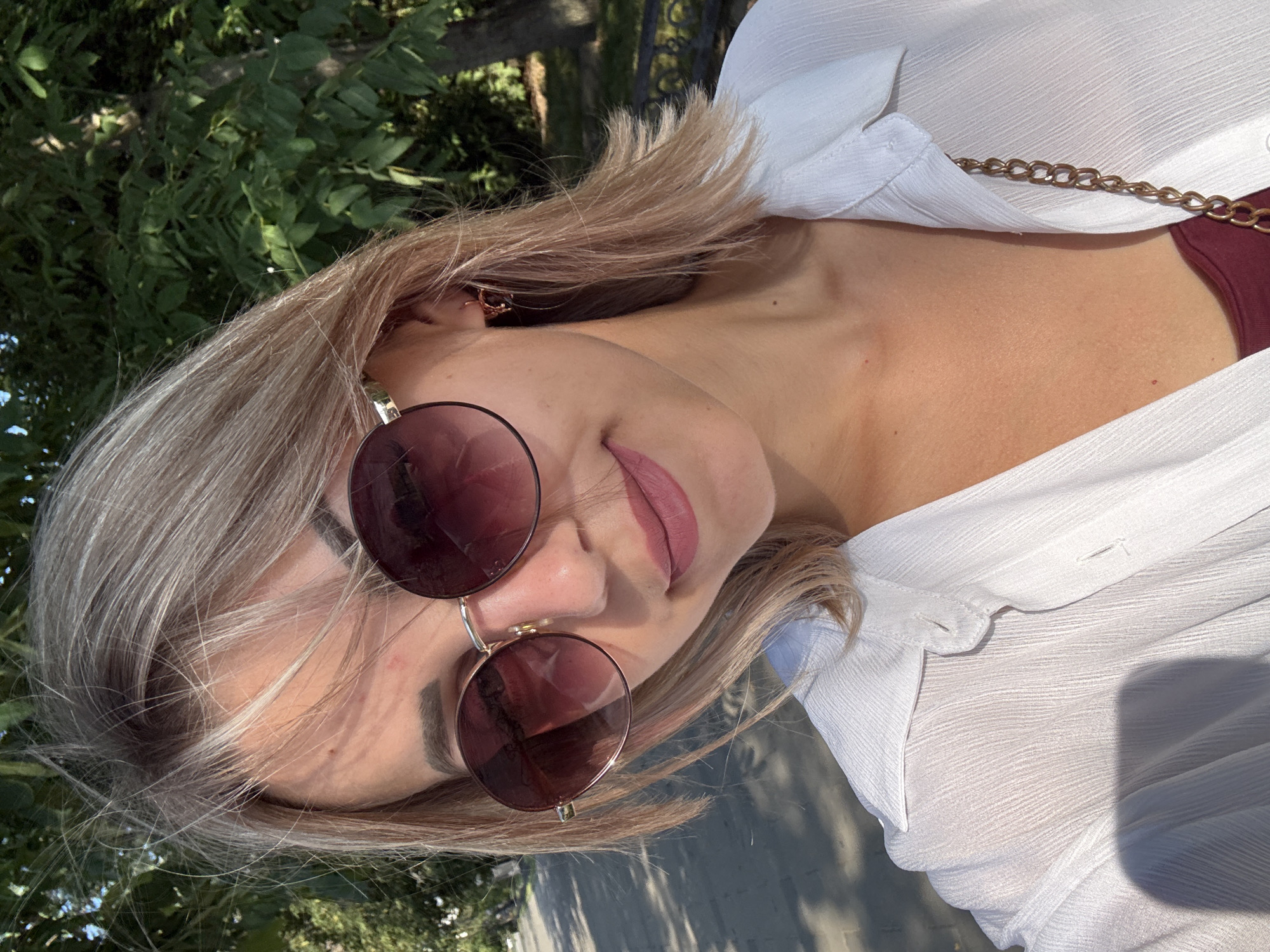 A young woman with shoulder-length blonde hair is posing for a selfie outdoors. She is wearing large, round, dark-lensed sunglasses and a white, semi-sheer collared shirt. A thin gold chain is visible over her shoulder. The background consists of lush green trees and foliage, with a paved pathway and dappled sunlight on the ground to her left. The overall scene suggests a sunny day in a park or outdoor recreational area. Based on the provided information, the location is Yuzhno-Sakhalinsk, Russia.